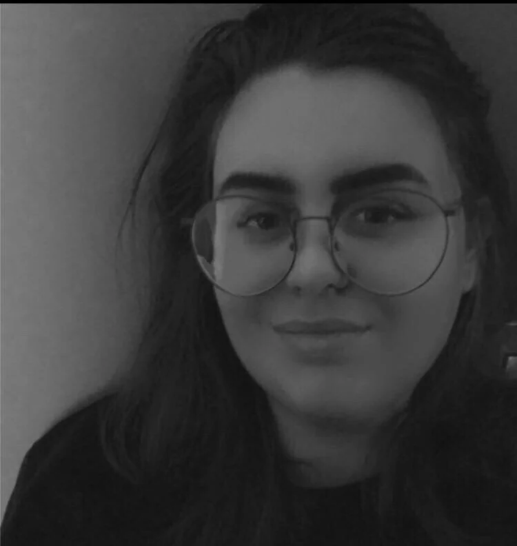 Black and white portrait of a woman wearing large round glasses, with dark hair parted to the side, looking at the camera with a slight smile.