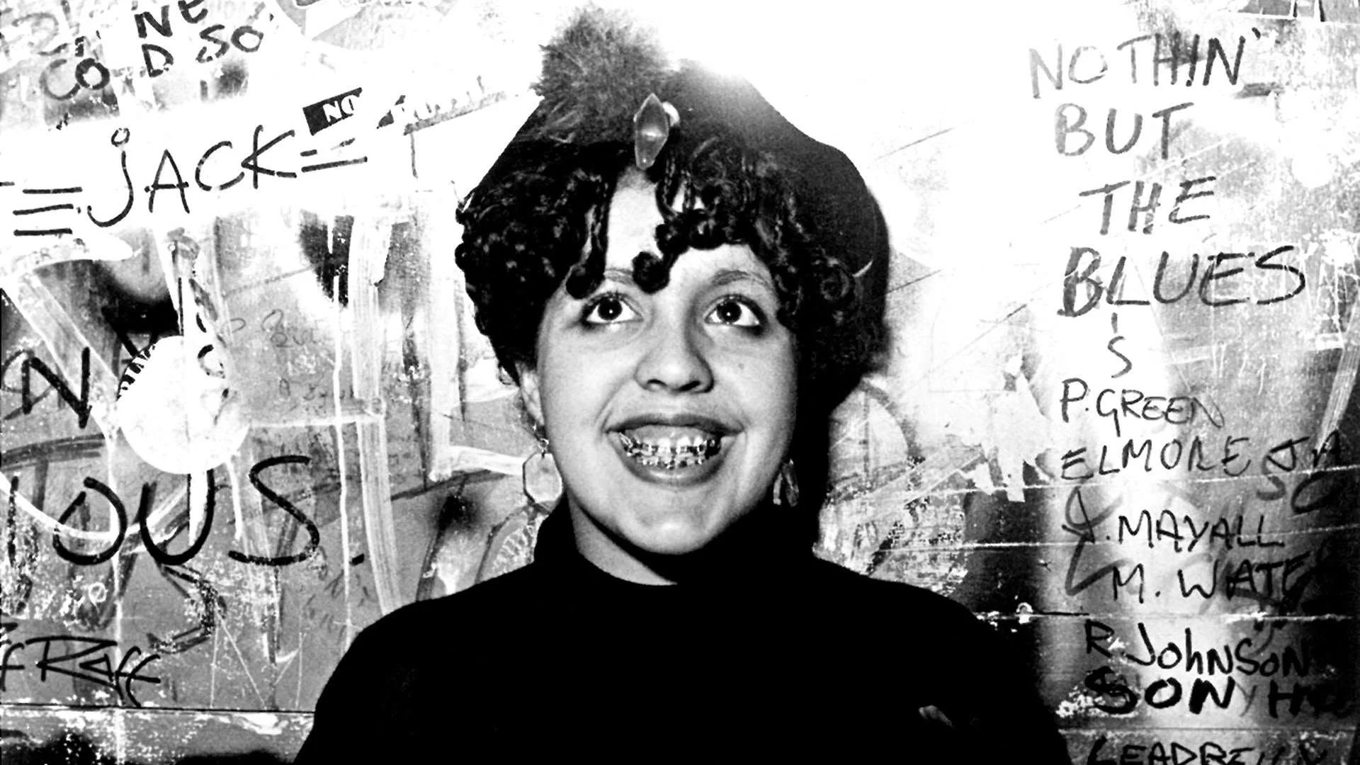 Black and white photo of Poly Styrene, a young mixed-race woman with short curly black hair, braces, and a black top, wearing a broad grin on her face.