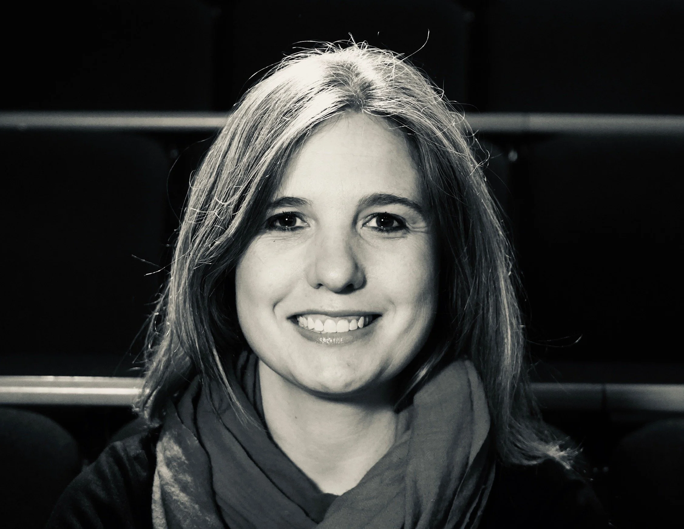 Black and white portrait of a woman smiling with shoulder-length hair, wearing a scarf and a dark top.