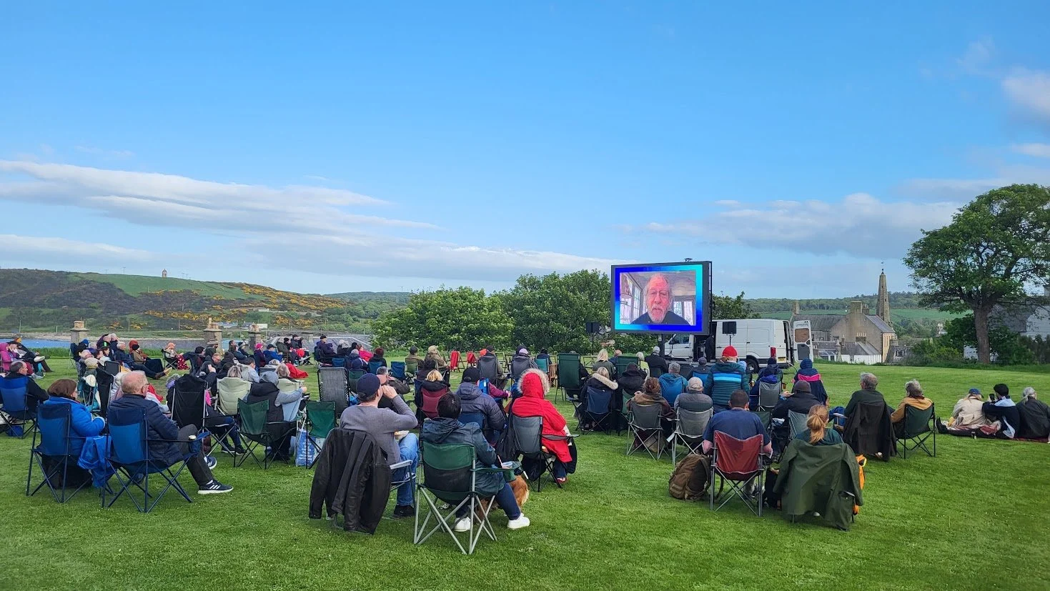 A large crowd watches actor Peter Riegert speak via zoom on a big screen, in a field overlooking the sea