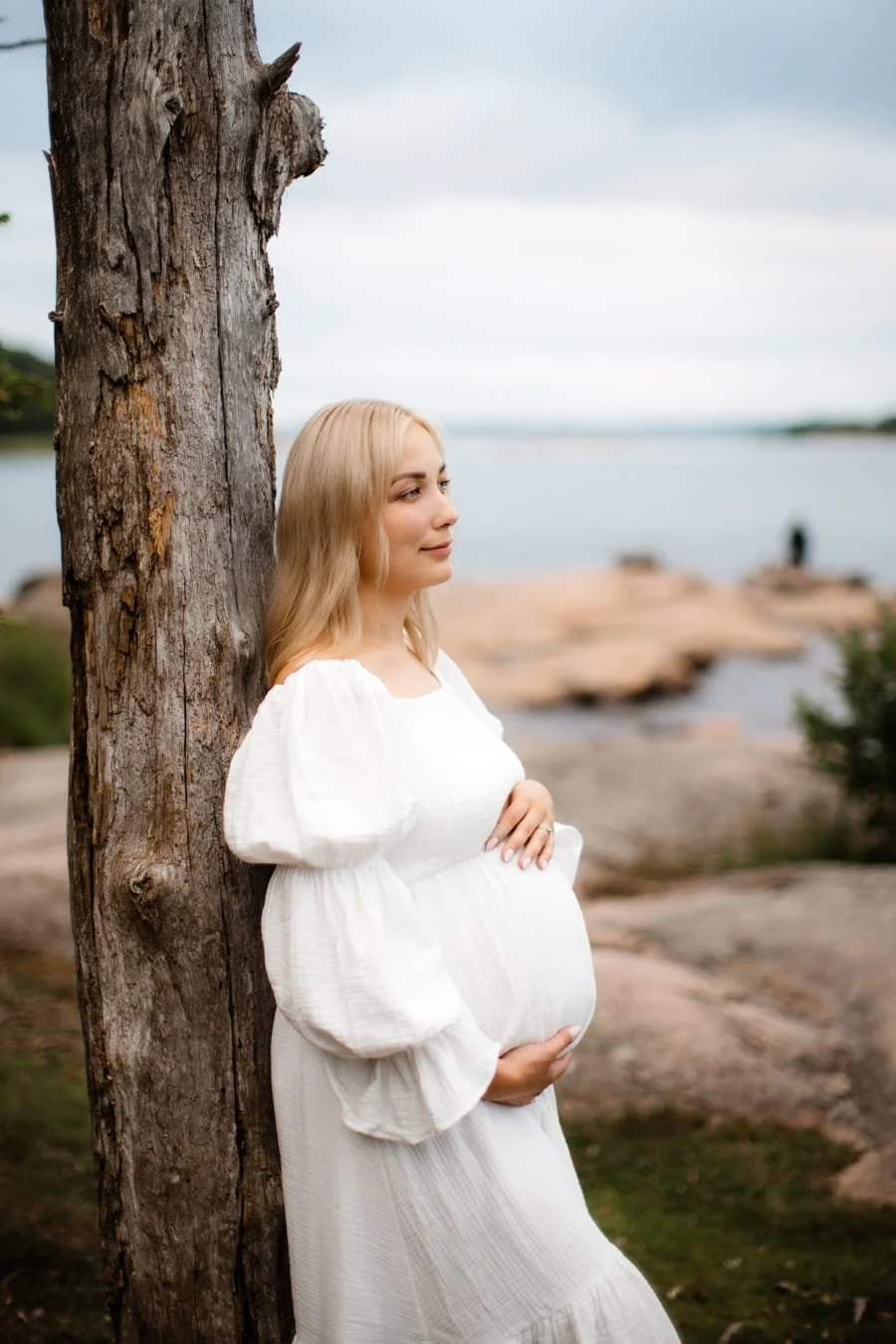 Raskaana oleva nainen nojaa puuhun rannalla, taustalla vesi ja pilvet.