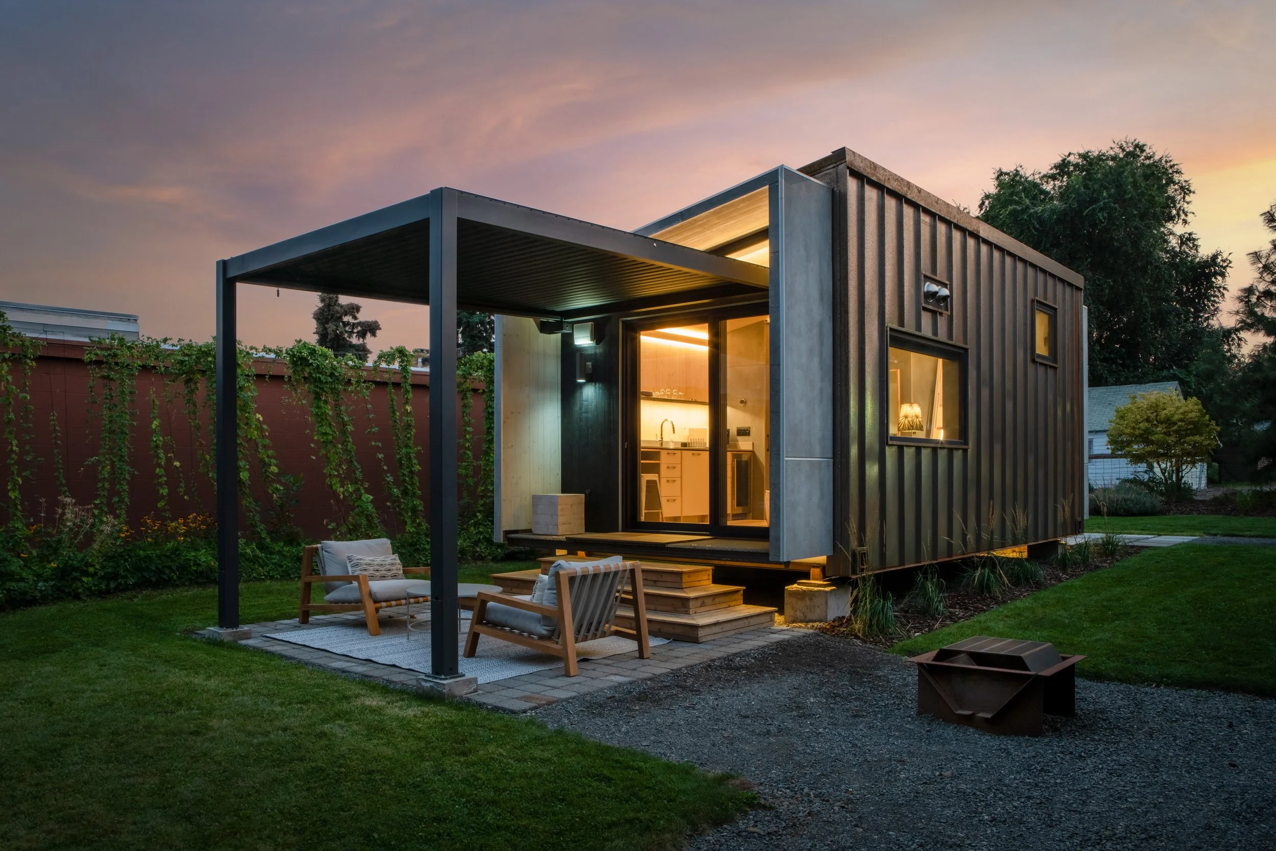 Modern tiny house with a porch and outdoor seating at sunset, surrounded by a lawn and trees.