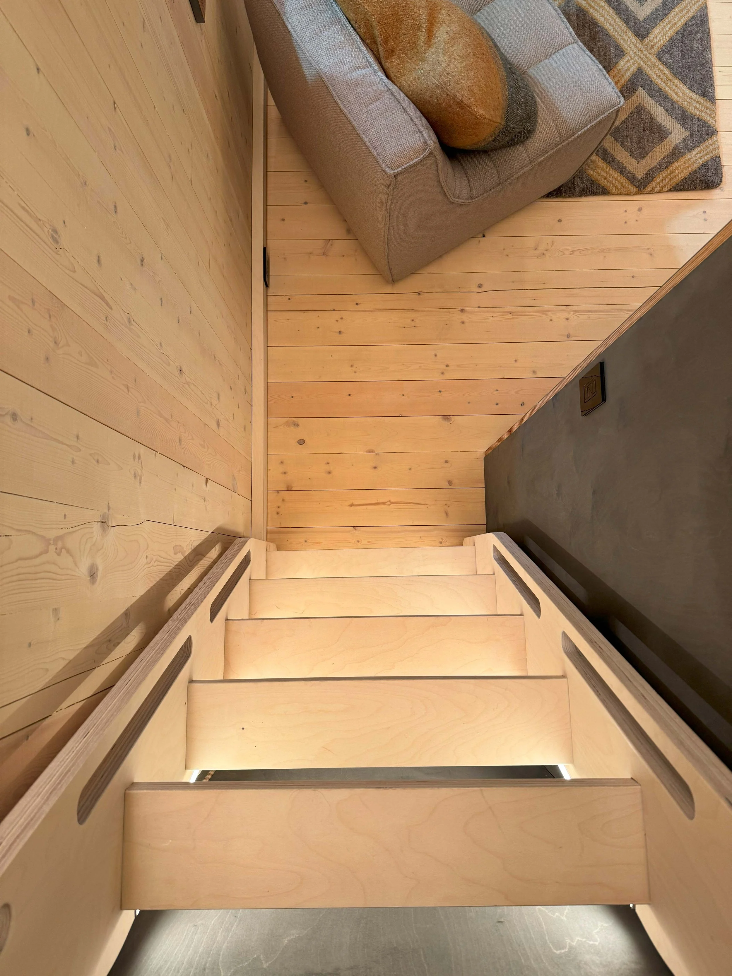 A wooden staircase with three steps leading up to a living room area with wood-paneled walls, a gray sofa with a decorative pillow, and a geometric patterned area rug.