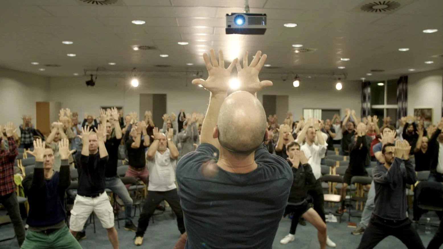 A facilitator raises his arms before a room of men at a masculinity camp, from Confessions of a Swedish Man