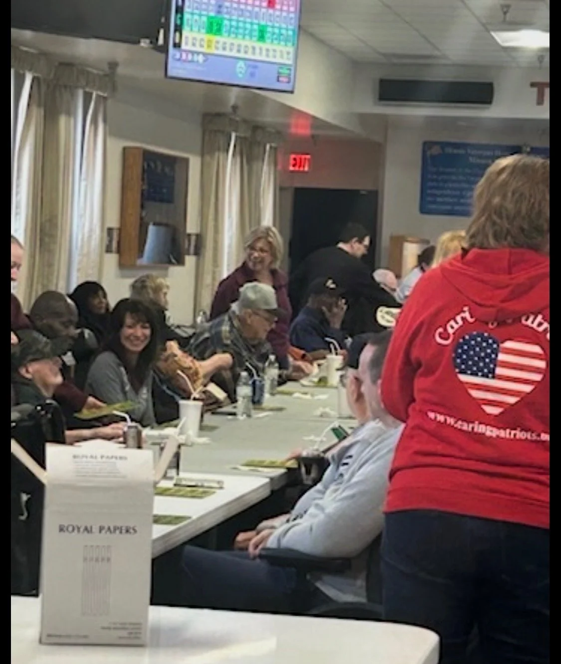 A group of people seated at a long table in a room with a person standing and smiling. Some are eating, and there are drinks and pamphlets on the table. A person in a red hoodie with a heart and American flag design is in the foreground.