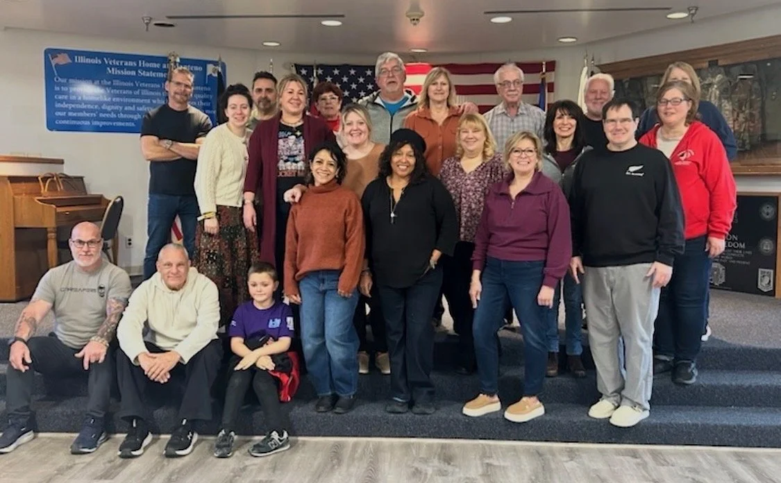 Group photo of people in a room with American flags and a blue sign about Illinois Veterans Home in the background.
