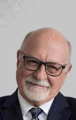 Portrait of an older man with glasses, white hair, and a beard, wearing a dark suit and tie, smiling against a plain background.