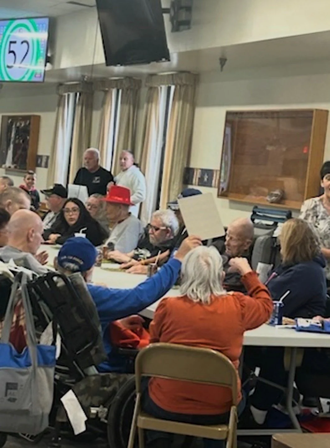 A group of diverse people of various ages sitting around a long table in a room with beige walls and curtains, some holding signs, with a few standing behind them. A screen displaying the number 52 and two men standing near a speaker in the background.