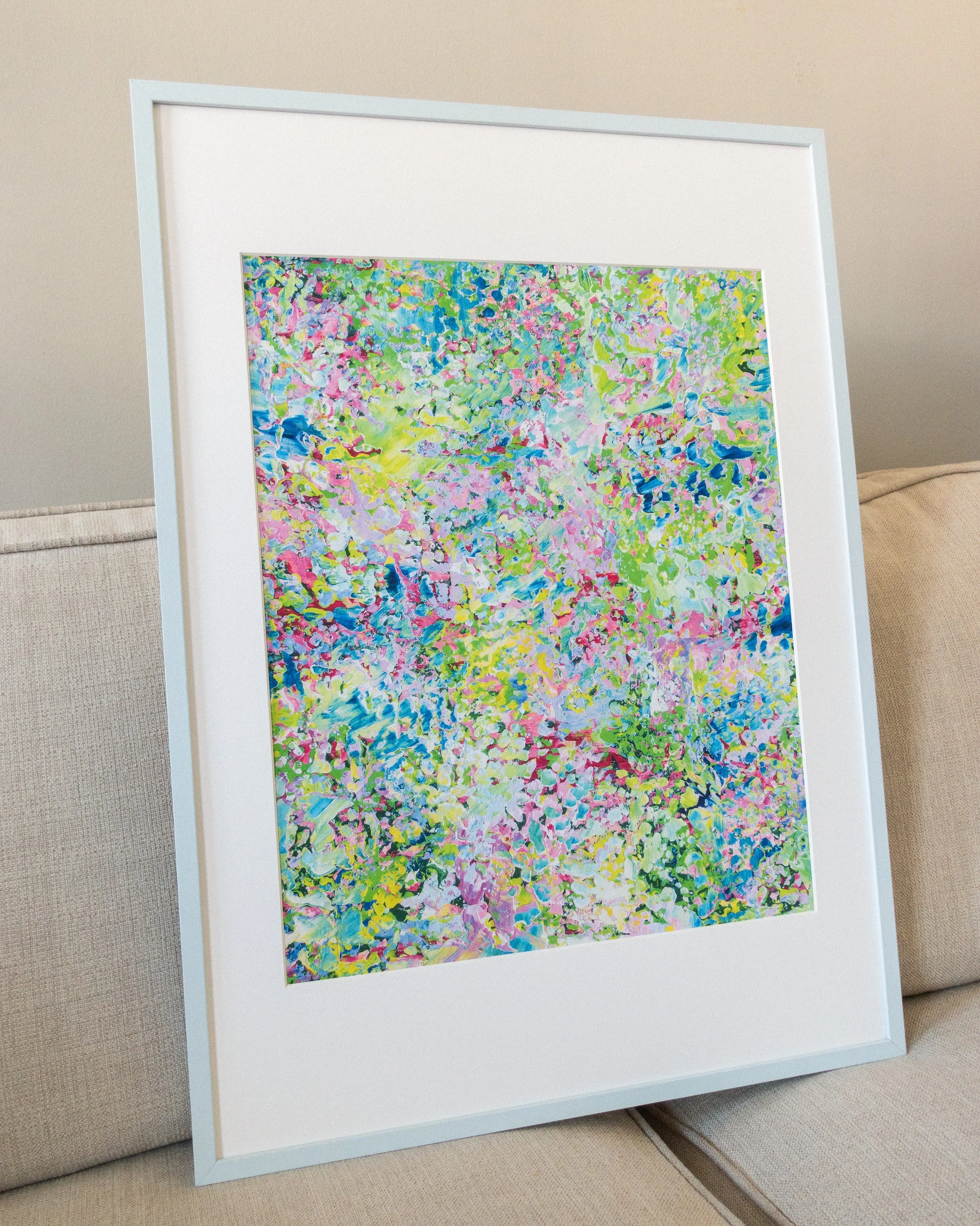 Inramad färgstark poster med passepartout på ljus soffa av Klara Collin med gröna, blå, rosa och lila detaljer. När knopparna brister poster från Klaras Kludd, inspirerad av vår och blommande färger.
