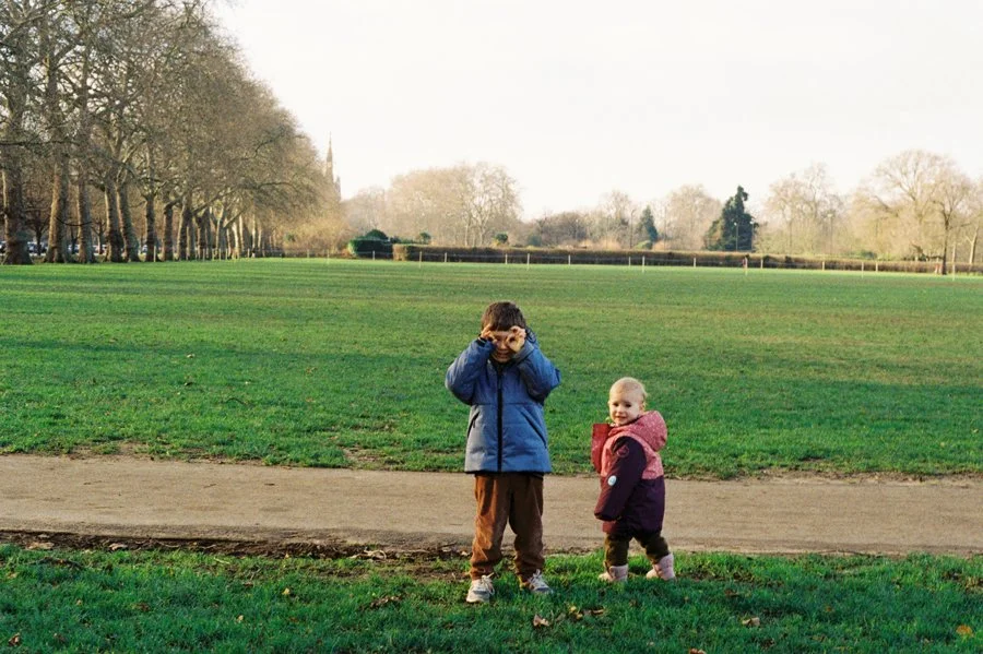 Two children standing in Hyde Park, London, on a cold winter afternoon. The boy makes pretend binoculars with his hands while his sister looks on. Photographed on film.