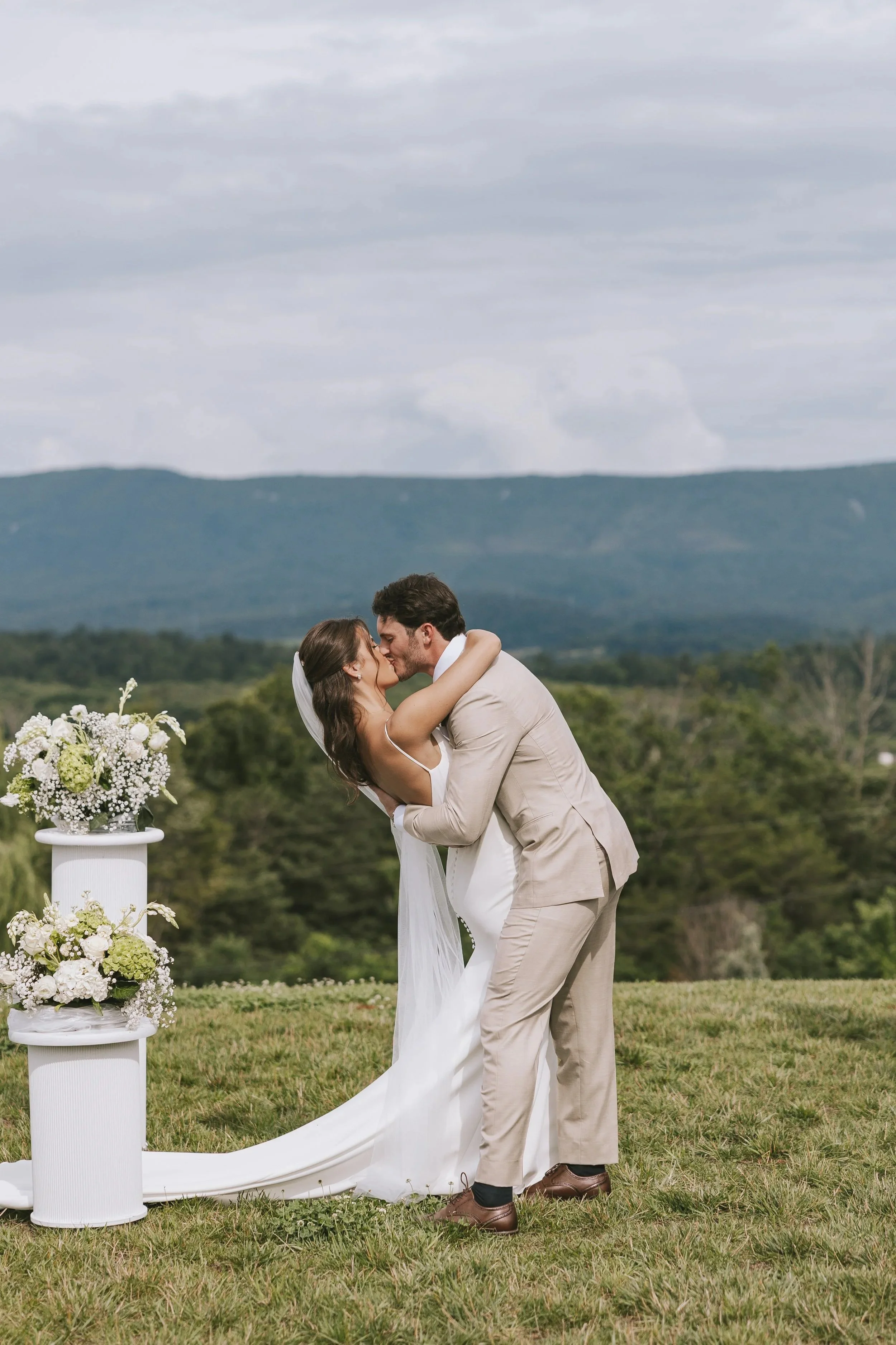 A newlywed couple kissing outdoors on their wedding day, with mountains and a cloudy sky in the background. The bride wears a white gown with a veil, and the groom is in a beige suit. There are white floral arrangements on white pedestals near them.
