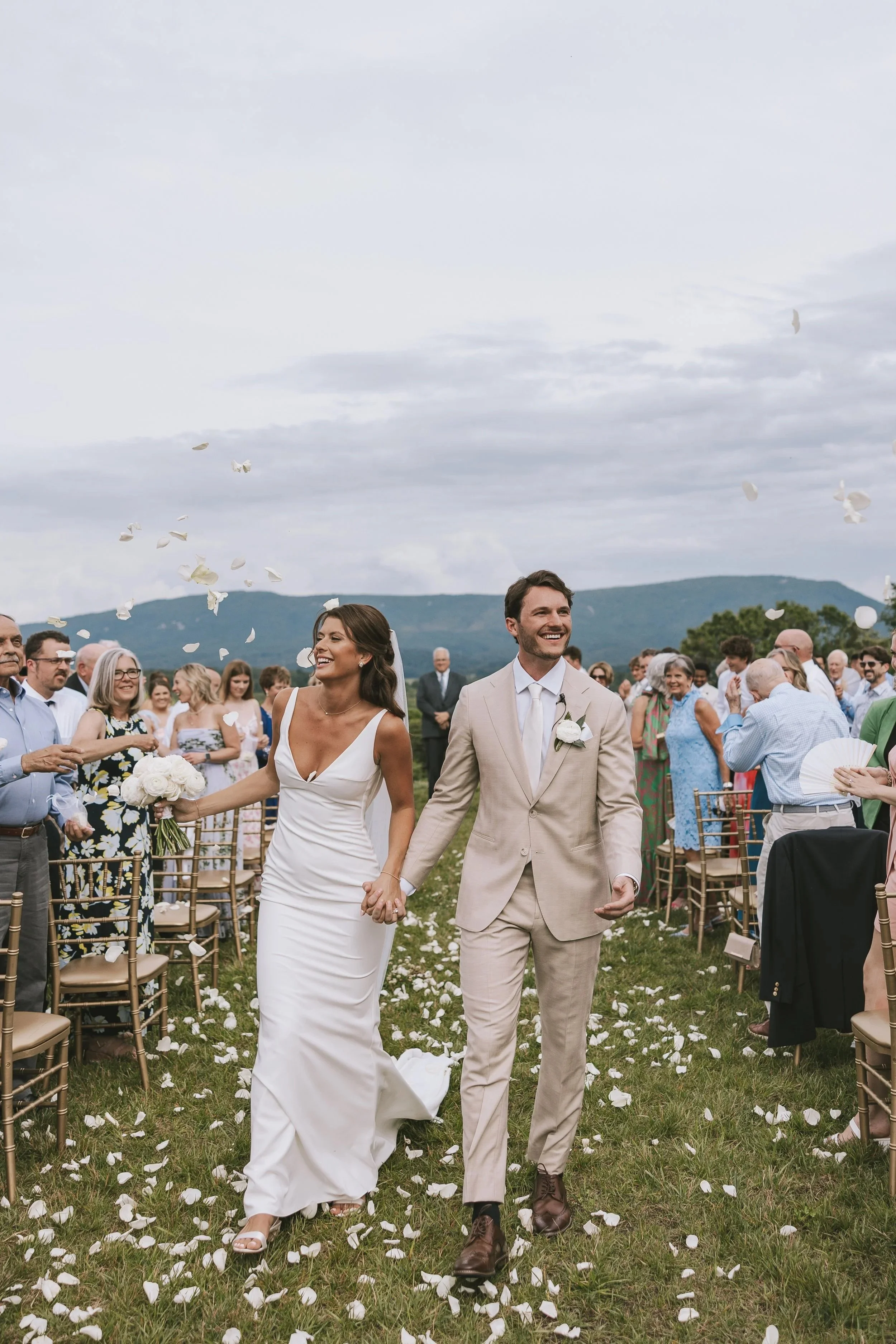 A bride and groom walking hand-in-hand down the aisle at an outdoor wedding ceremony at The James Morgan, surrounded by guests who are celebrating and throwing flower petals, with Blue Ridge mountains in the background.
