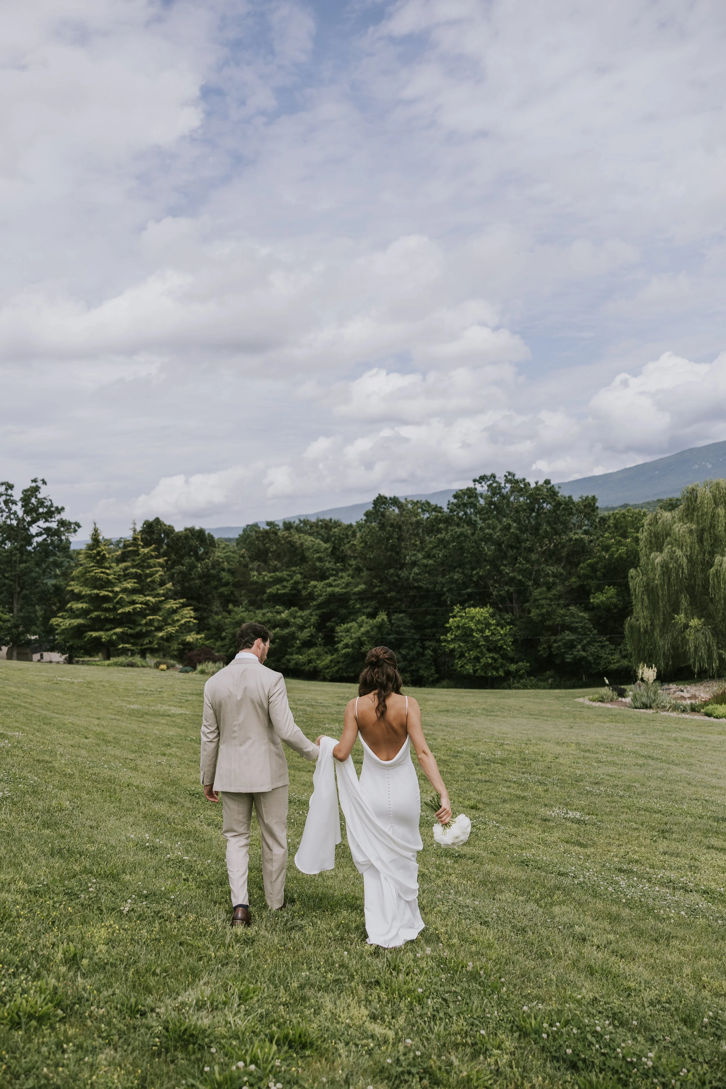 Bride and groom walking across a grassy field, holding hands, with the bride carrying a bouquet of white flowers and wearing a white wedding gown, and the groom in a light-colored suit, against a backdrop of trees and a partly cloudy sky.