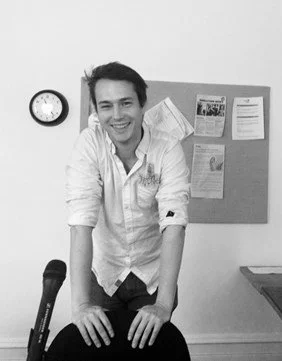 Éric Bordeleau is wearing a white shirt and leans on a chair with two hands while smiling. There is a microphone in front of him. In the background is a bulletin board and clock.