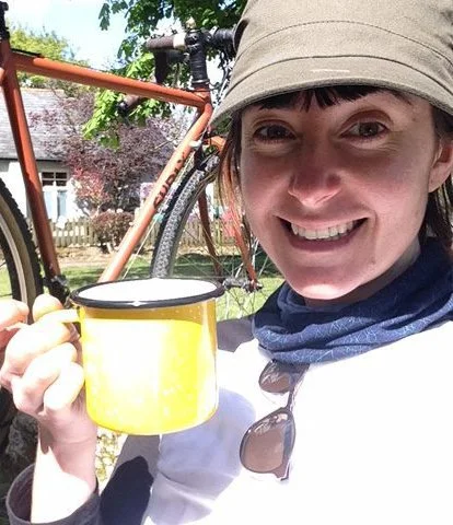 Becky wears a white t-shirt, an olive hat, and blue scarf. She is smiling and holds a yellow mug in her right hand. In the background there is a bicycle and cottage.