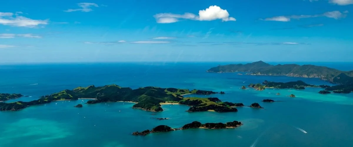 Oceanic connections - A photo of the Bay of Islands in Aotearoa New Zealand. The Māori name - Ipipiri - translates to ‘many places’. The photo shows green islands spread throughout the bay with a clear blue sky in the background and a few white cloud