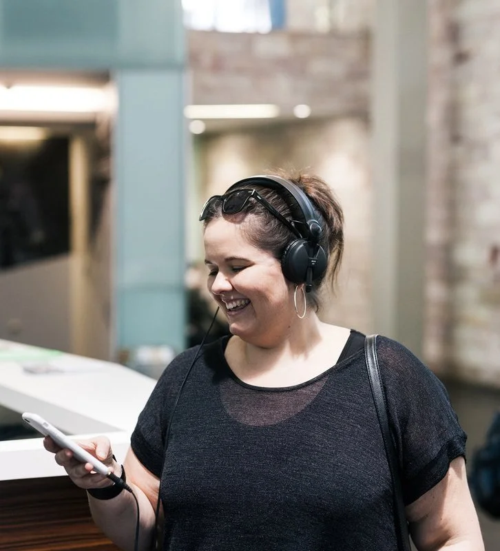 Leah Barclay is wearing a black shirt with a black bag strap over her shoulder, and also black headphones. She smiles while looking down at her phone. The background is blurry.