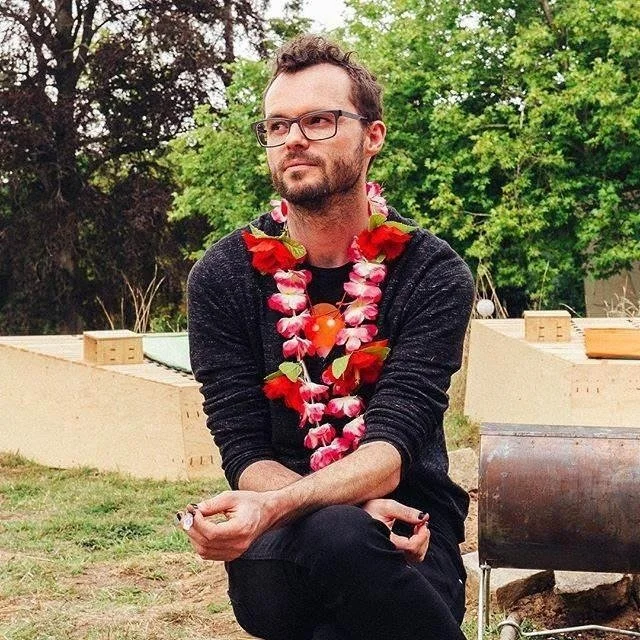Mix Irving is wearing a black shirt, black-rimmed glasses and matching black nail polish. Mix is garlanded in a fresh flower lei with pink and red flowers. In the background there are green and brown leafy trees and a wooden workshop.