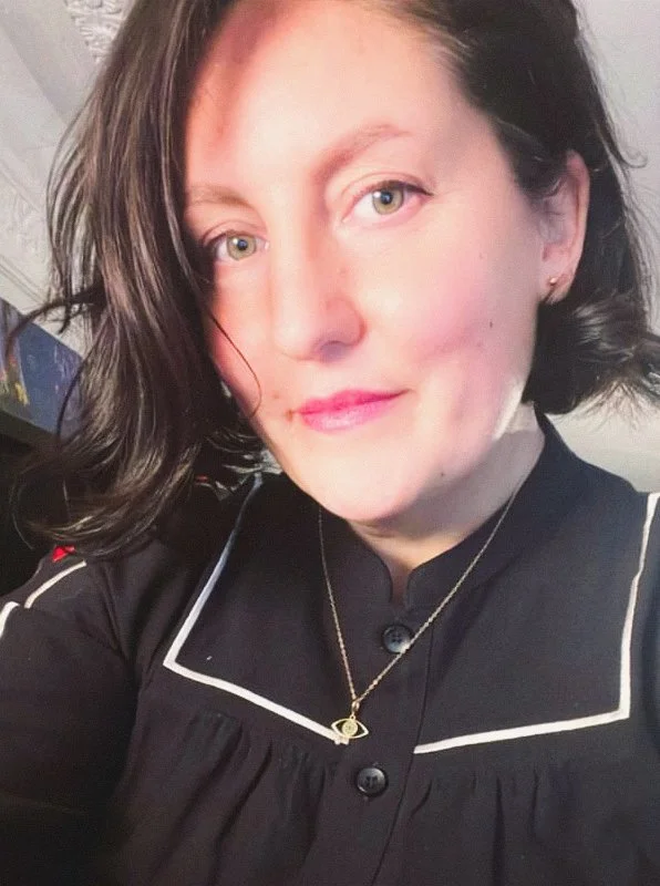 Cassie is wearing a black top with white trim and a necklace with an eye pendant. She is smiling at the camera and wears pink lipstick.