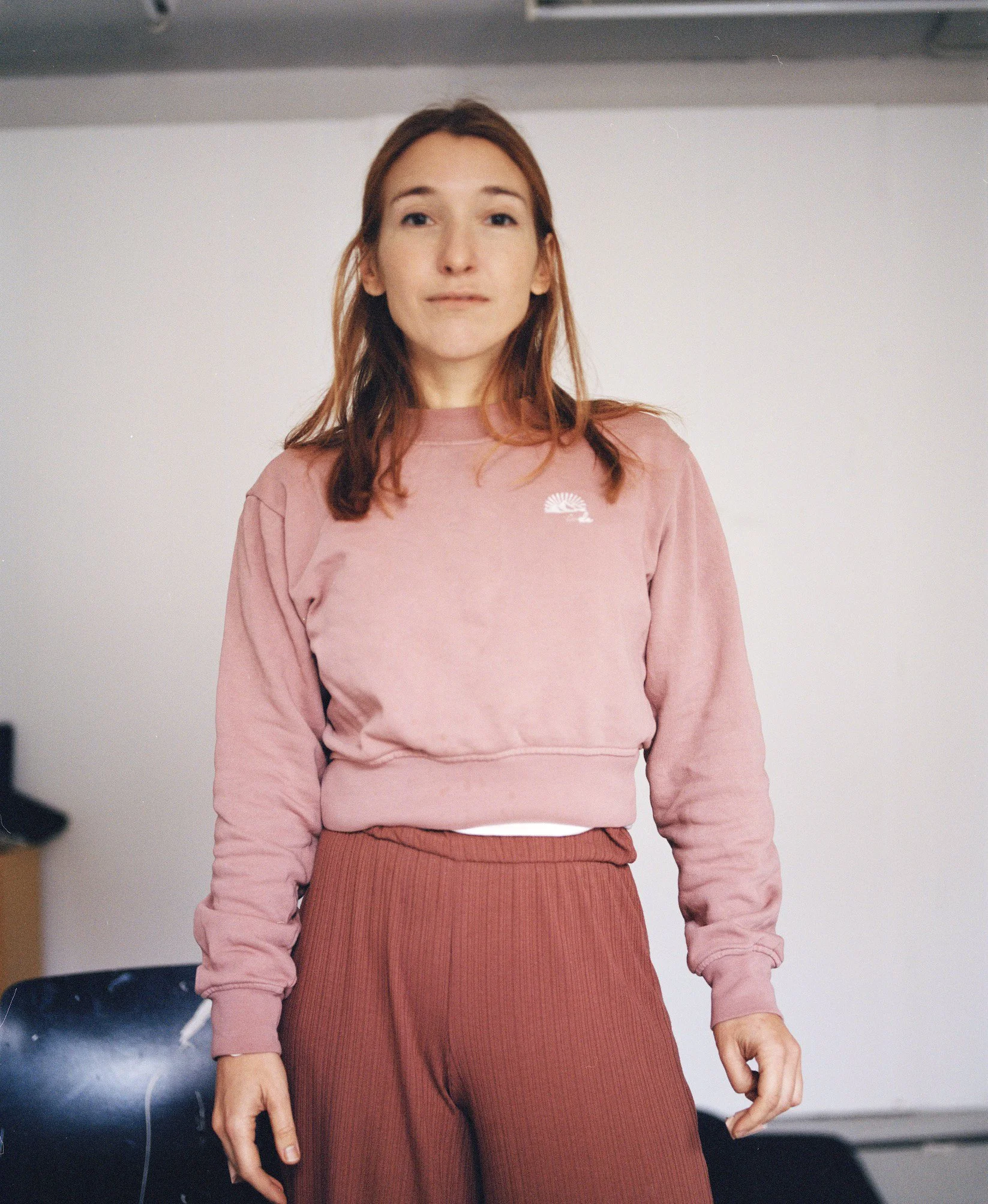 Lene smiles and is facing the camera. She wears a pink pullover and loose trousers. In the background, the white wall of the dance studio.