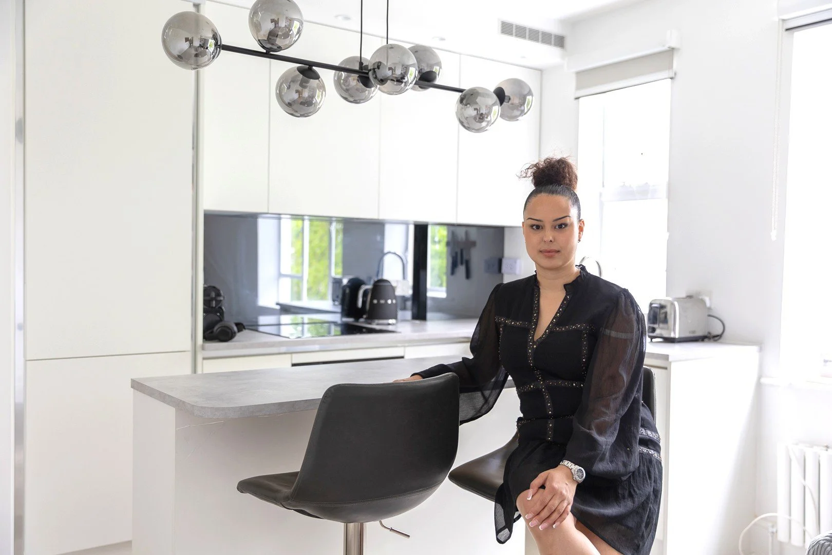 A woman with dark hair in a bun sitting at a kitchen counter in a modern white kitchen with black accents and large windows.