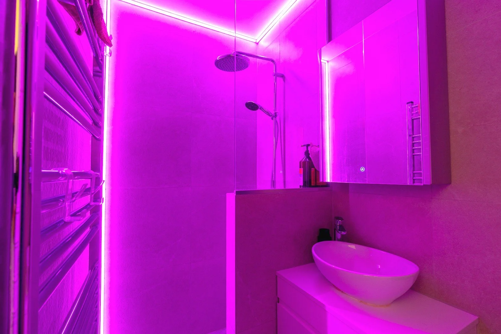 A bathroom illuminated with vibrant pink or purple lighting, with a shower area featuring a rain shower head and a handheld shower, a mirrored cabinet, a sink with a vessel basin, and a wall-mounted towel rack.