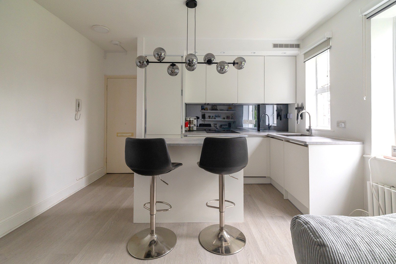 Minimalist kitchen with white cabinets, black bar stools, and a modern black chandelier, brightened by natural light from large windows.
