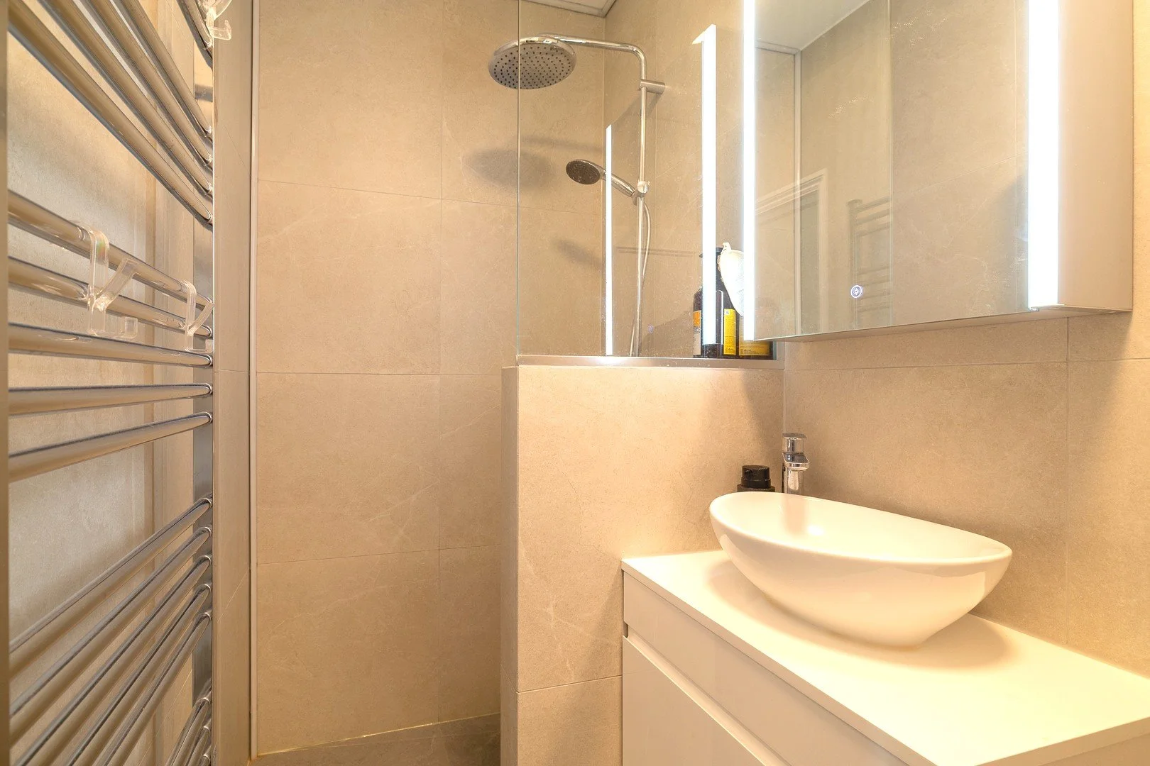 A modern bathroom with beige tiled walls, a walk-in shower with a rain shower head, a mirrored cabinet with LED lighting, a white vessel sink on a white vanity, and a heated towel rail on the left.
