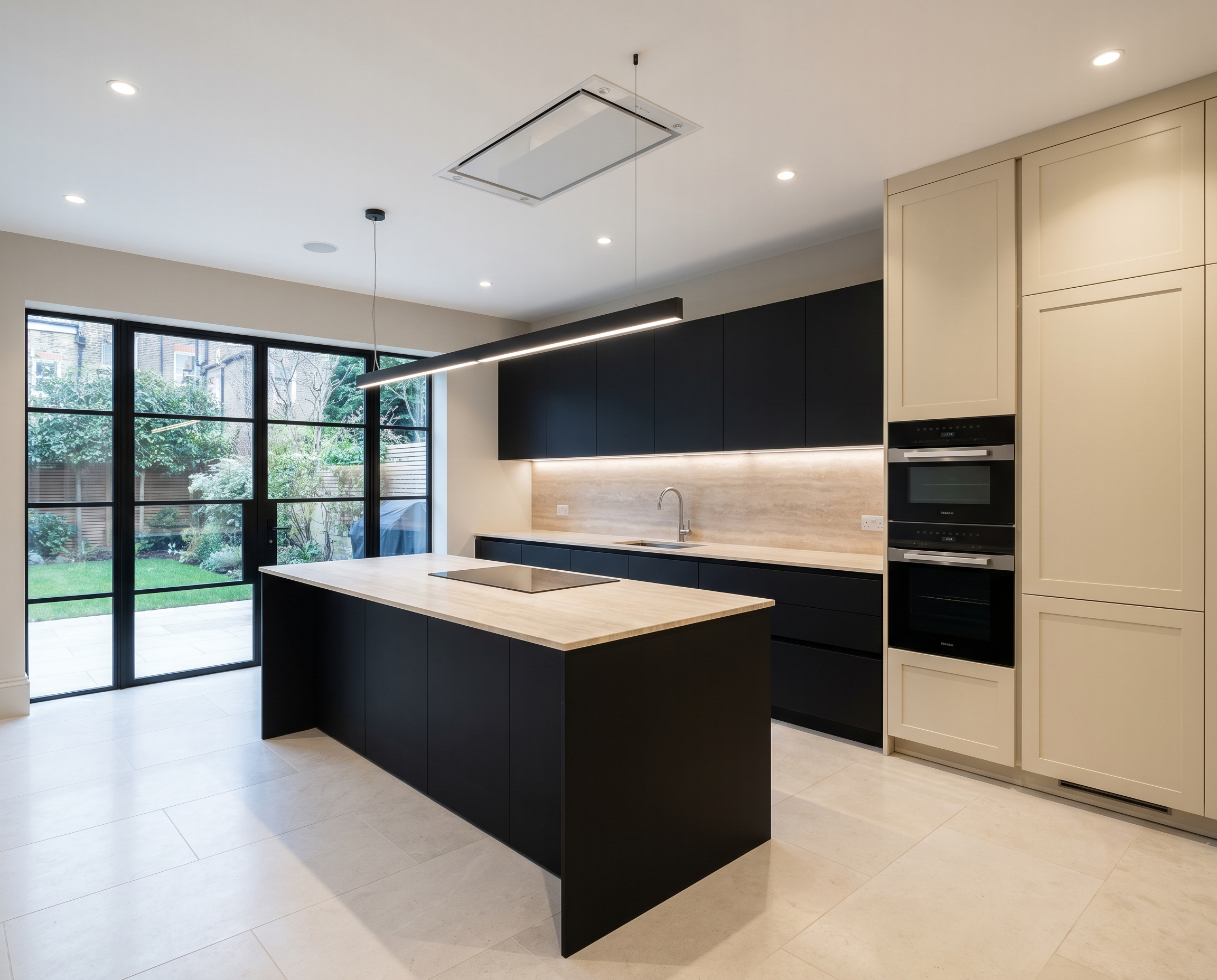 Modern kitchen with black and beige cabinets, island with light wood countertop, large windows showing a backyard, and ceiling lights.