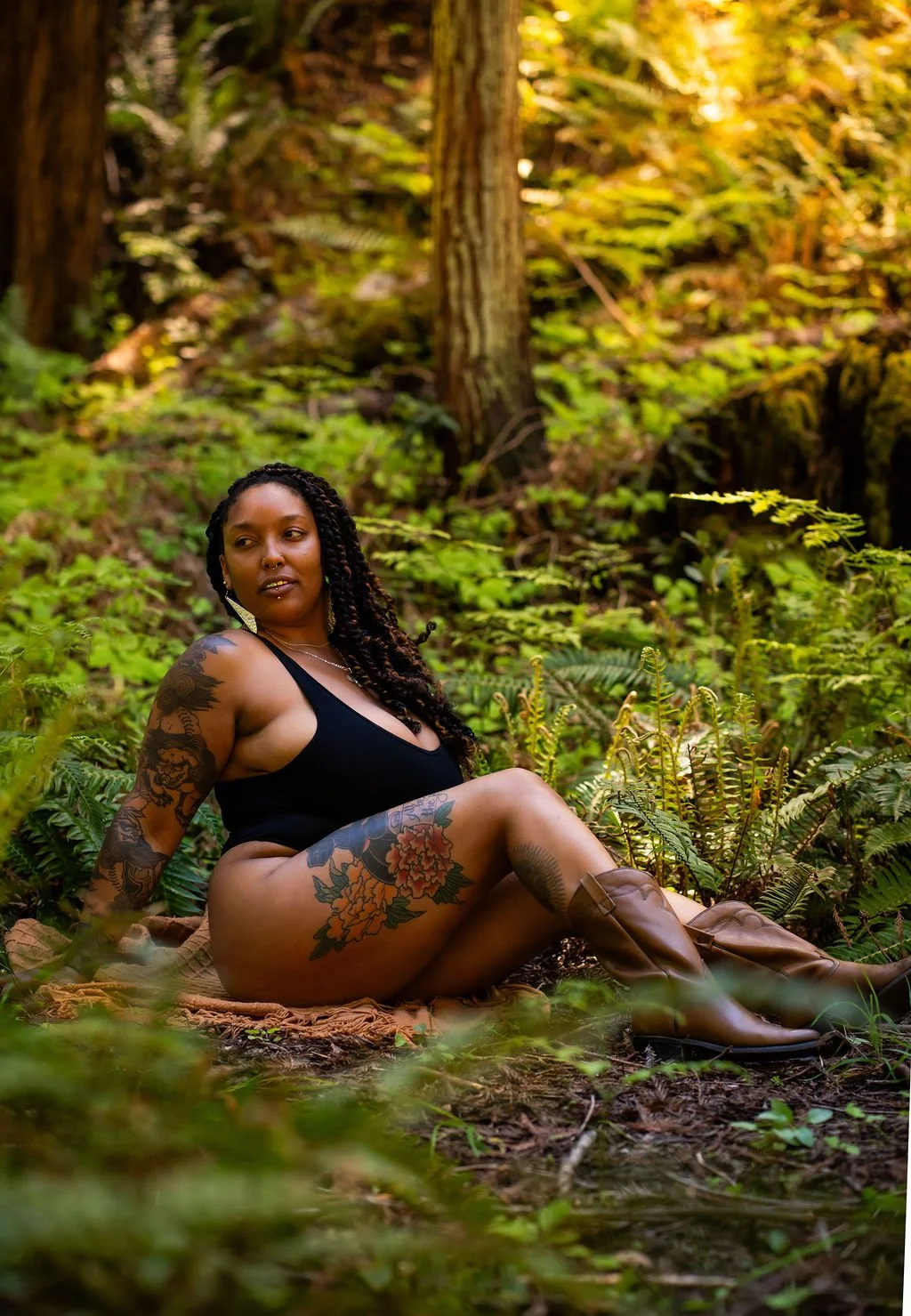 A tattooed Black woman wearing a rainbow opal gold grill and a black bodysuit sits on an orange blanket in a forest as she looks off into the distance.