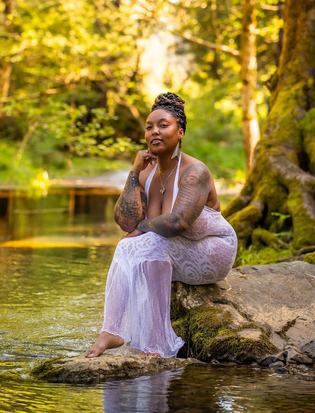A Black woman with tattoos and facial piercings wears a white lace dress as she sits on a rock next to a lake as she smiles at the camera.