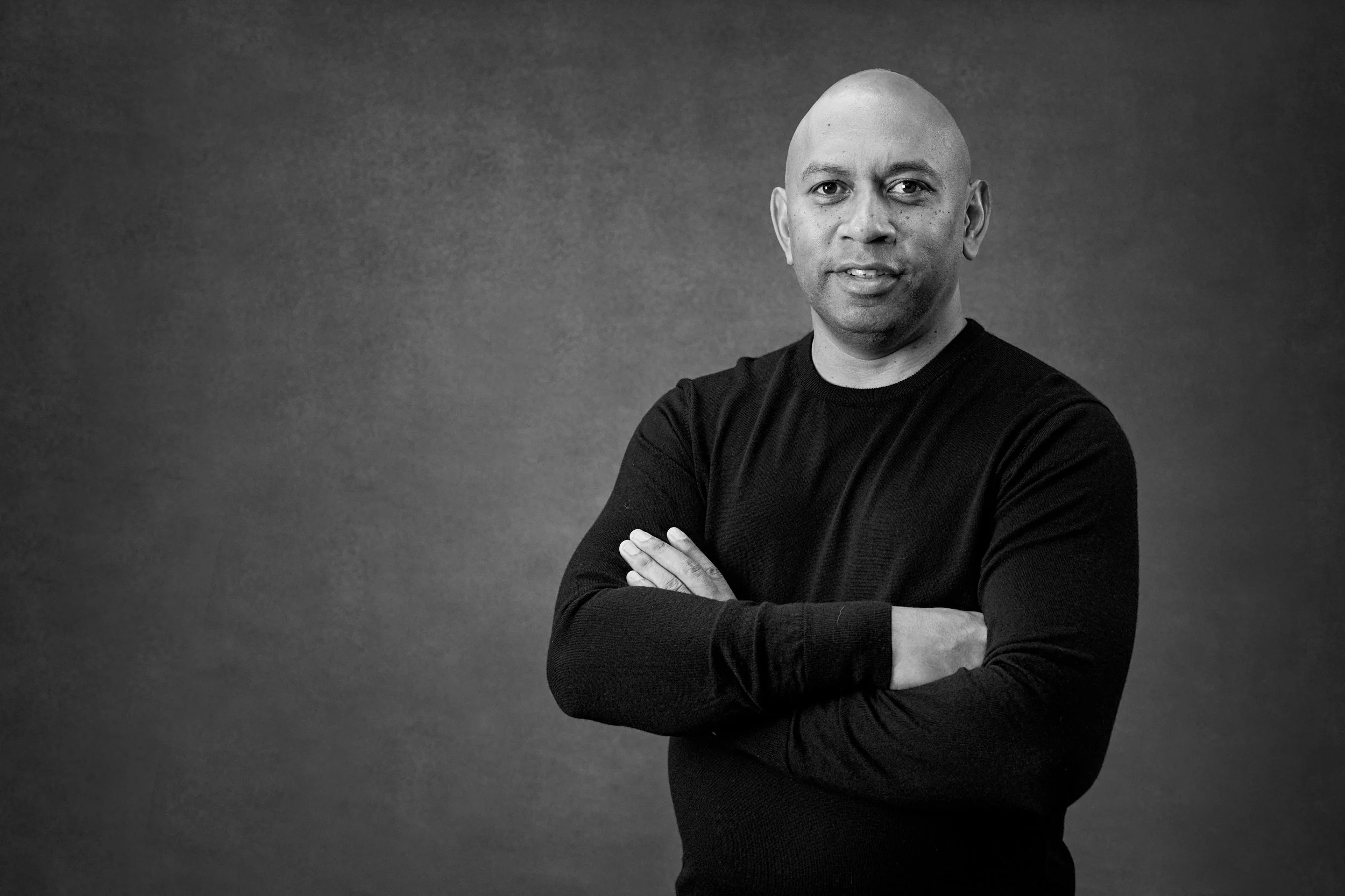 A black and white portrait of a bald man with light skin, wearing a black long-sleeved shirt, standing with arms crossed against a plain background.