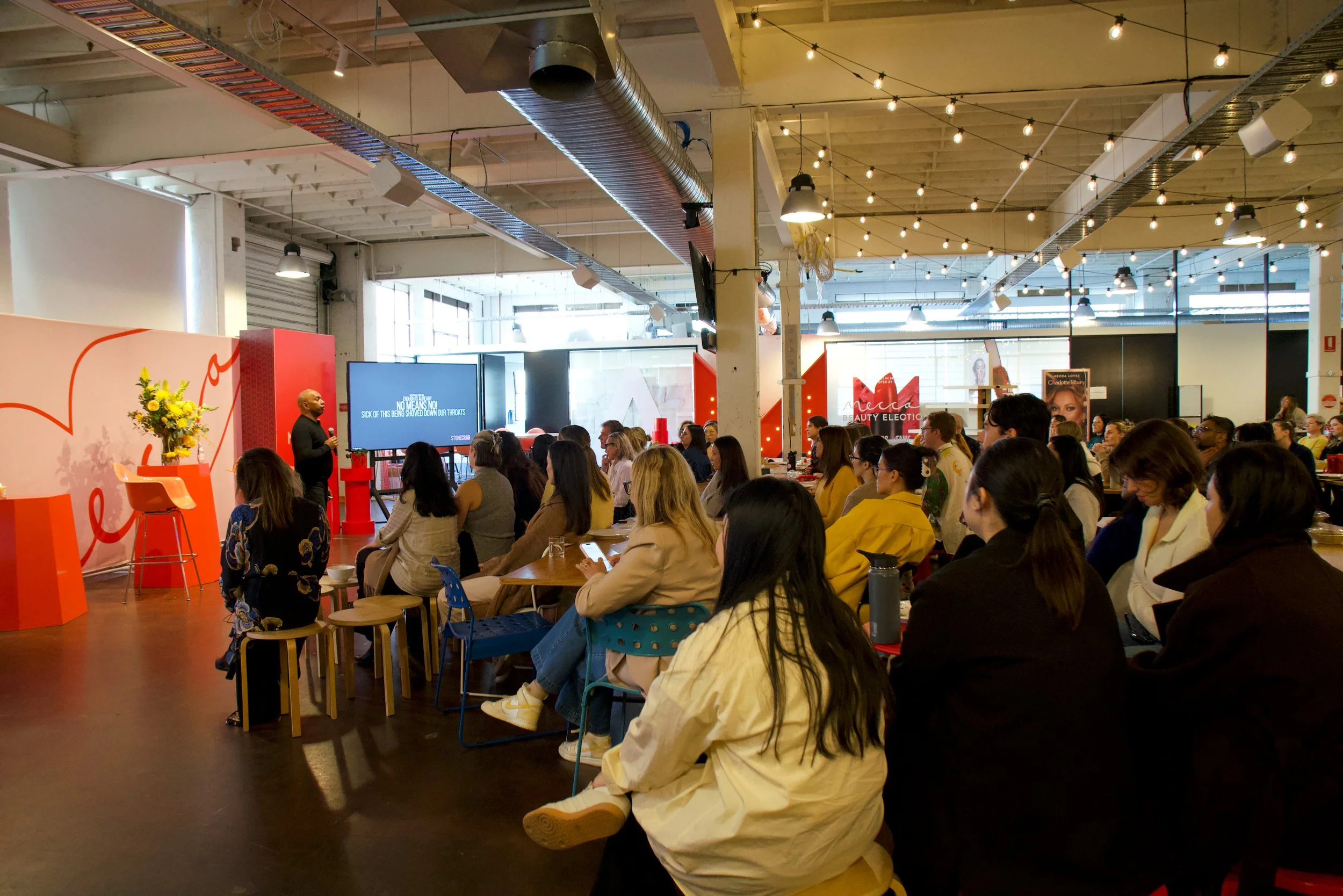 A large crowd of people seated at tables attending a presentation or event in an industrial-style venue with exposed ceiling ducts, hanging lights, and a large screen displaying text.