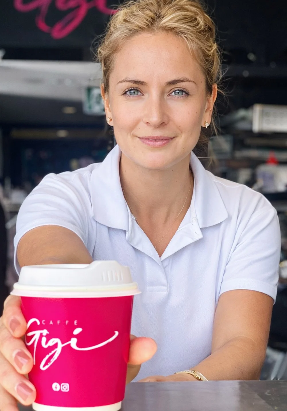 Caffè Gigi branded coffee cup mockup featuring a staff member holding a bright pink takeaway cup with white logo in a café setting.