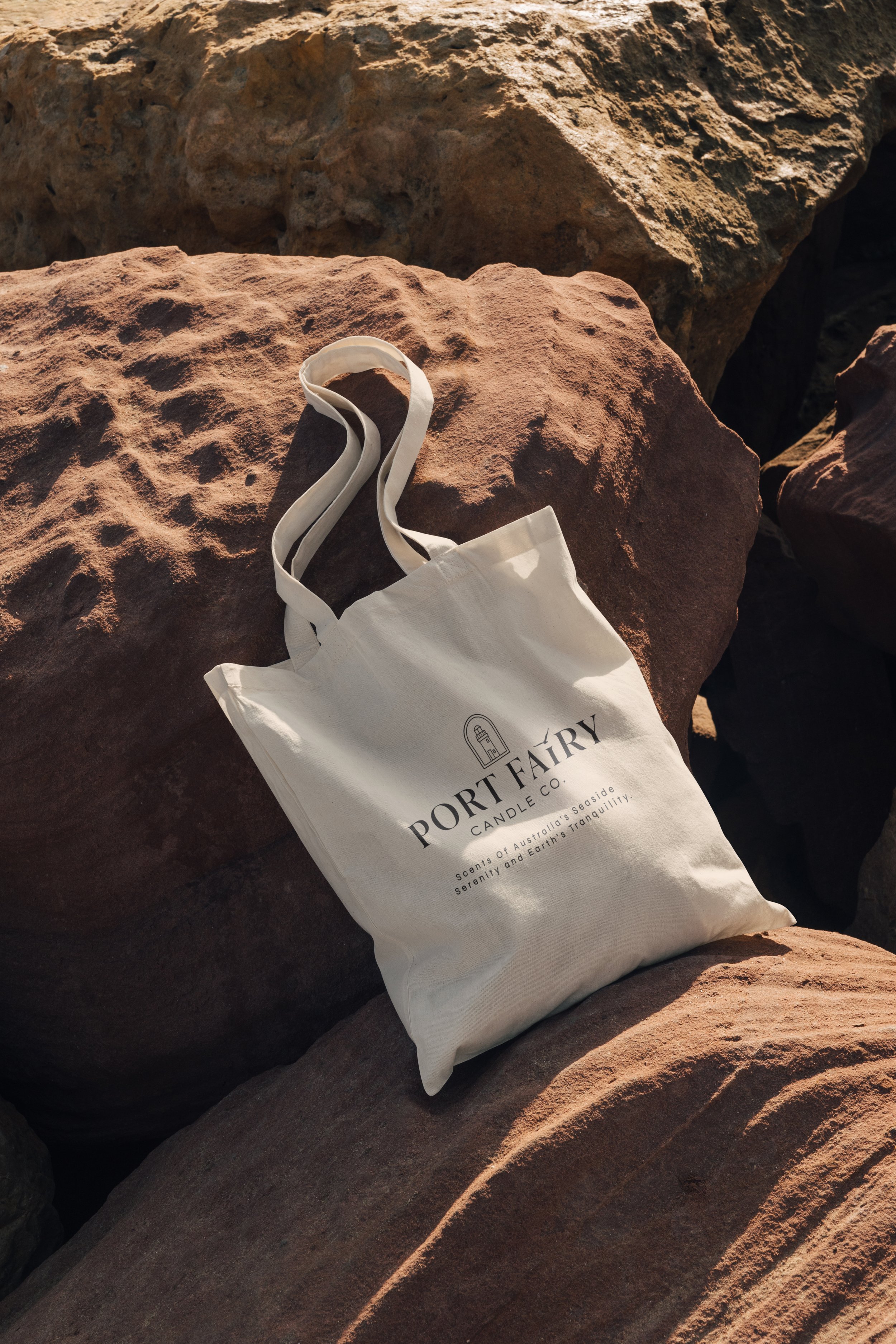 Port Fairy Candle Co. tote bag mockup featuring a branded cream canvas bag styled on coastal rocks with natural Australian seaside branding.