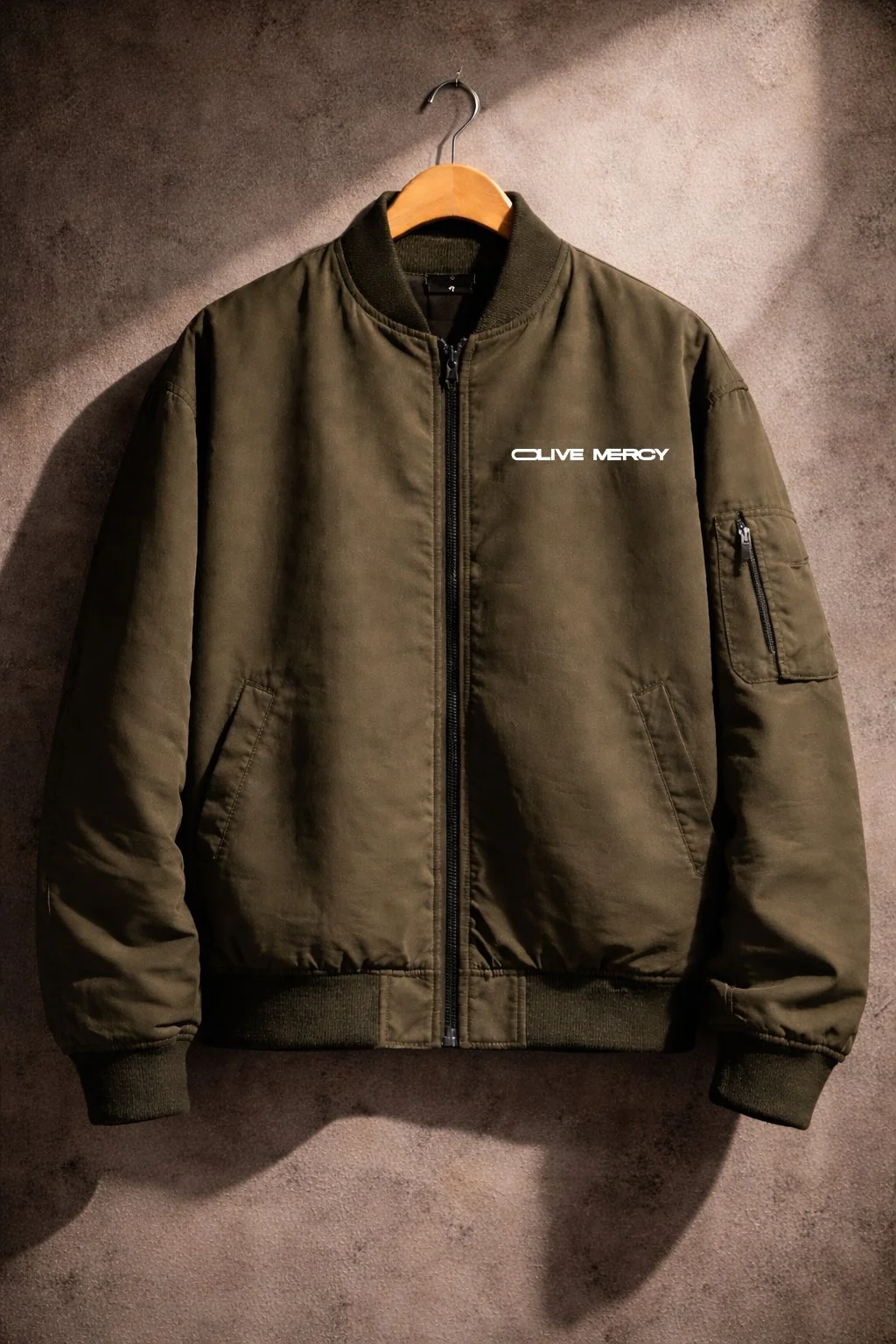 Olive Mercy olive bomber jacket mockup featuring a minimal white logo on a textured wall background.