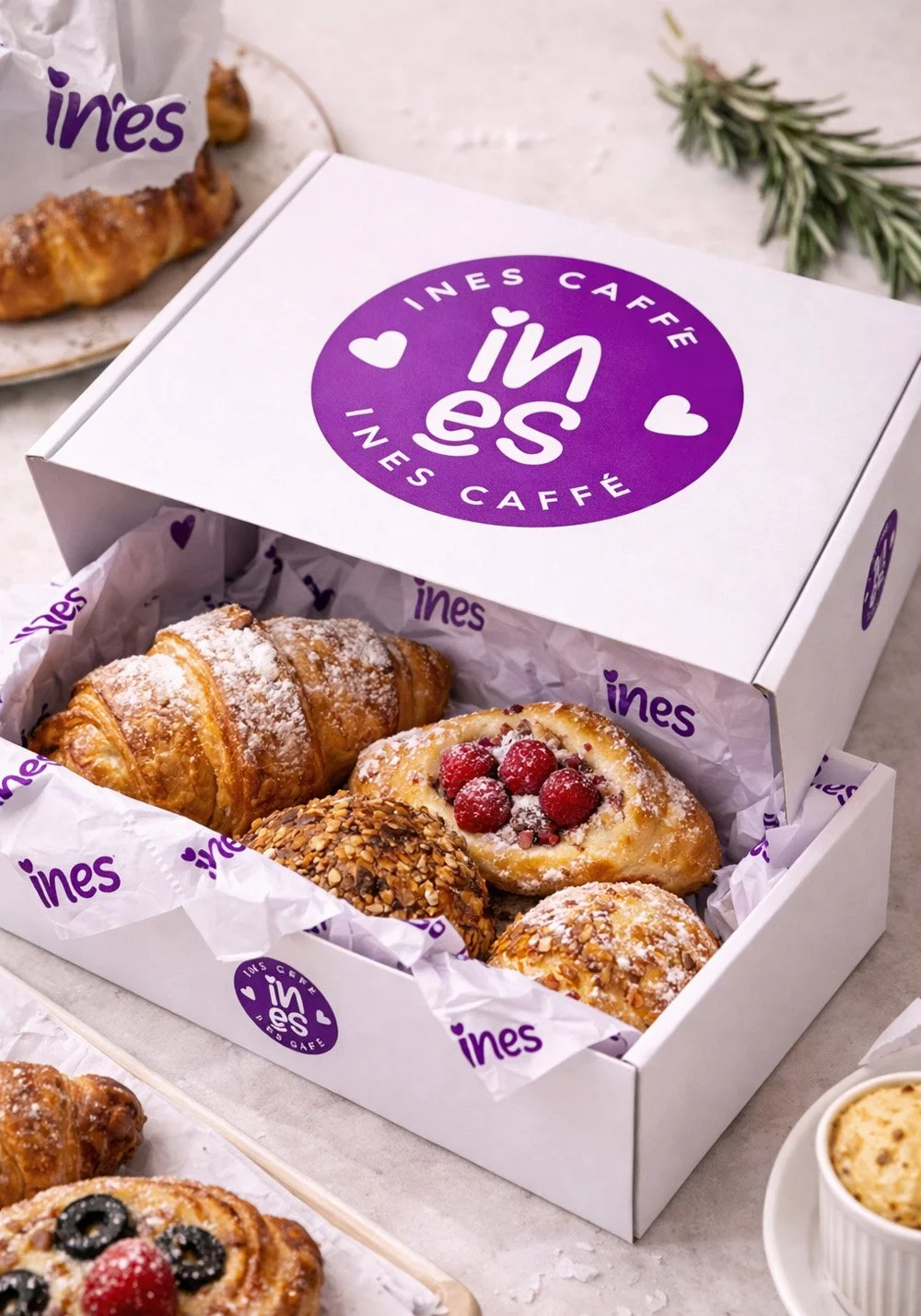 Ines Caffe pastry box packaging mockup featuring branded white box, purple logo, custom tissue paper and assorted pastries in a café setting.