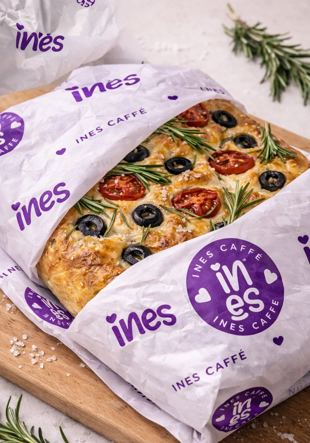 Ines Caffe focaccia wrapping paper mockup featuring branded purple tissue paper, café logo and fresh focaccia with rosemary, olives and tomatoes.