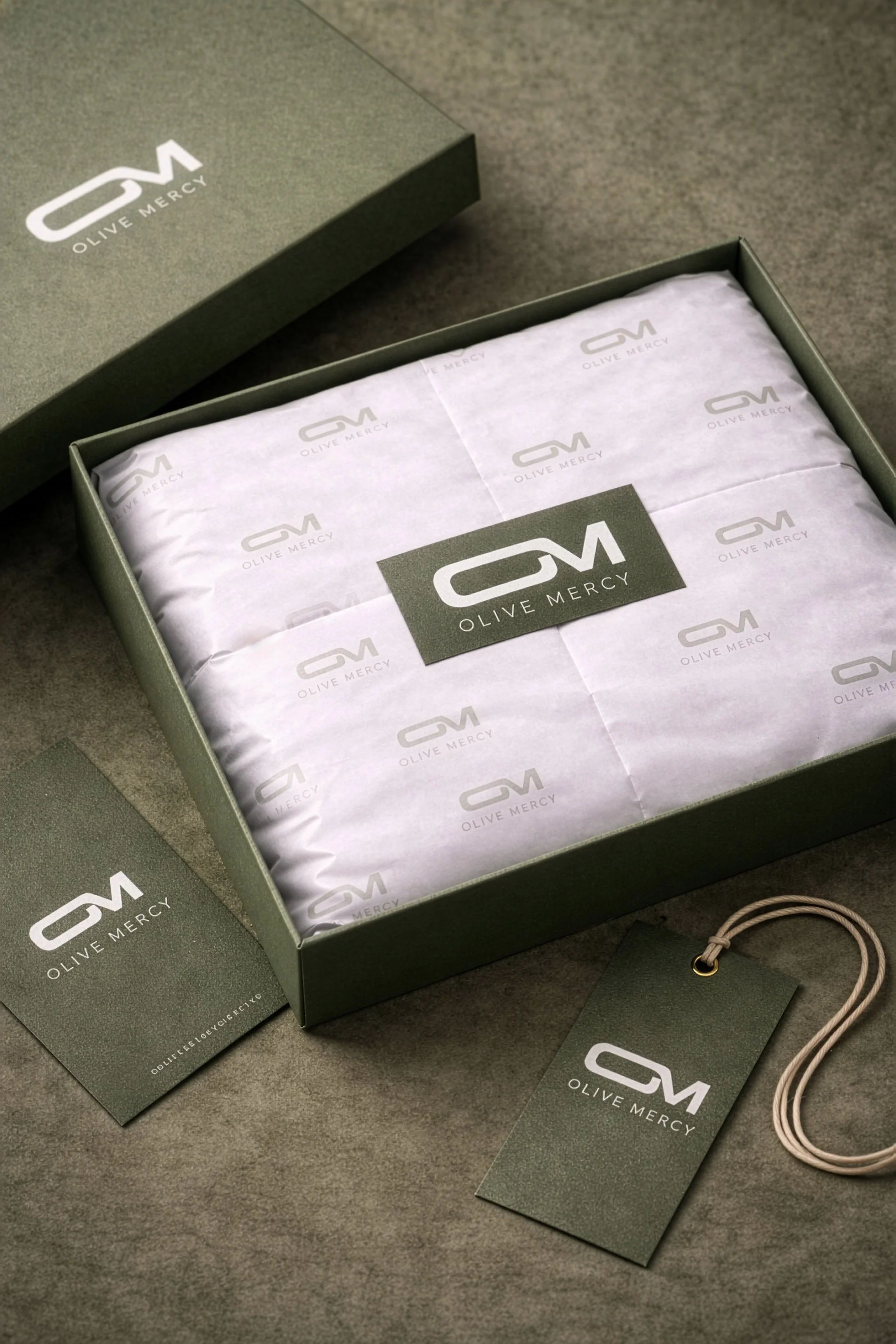 Olive Mercy olive green packaging mockup featuring a branded box, tissue paper, swing tags and OM logo details.