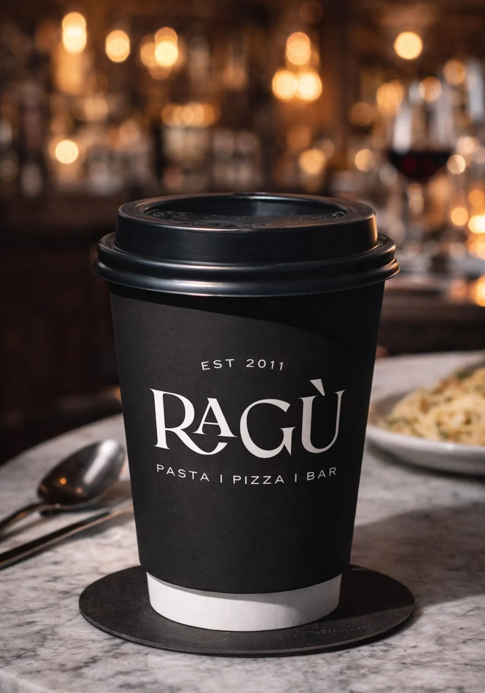 Ragù Pasta Pizza Bar branded black takeaway coffee cup mockup featuring the white logo in a moody Italian restaurant setting.