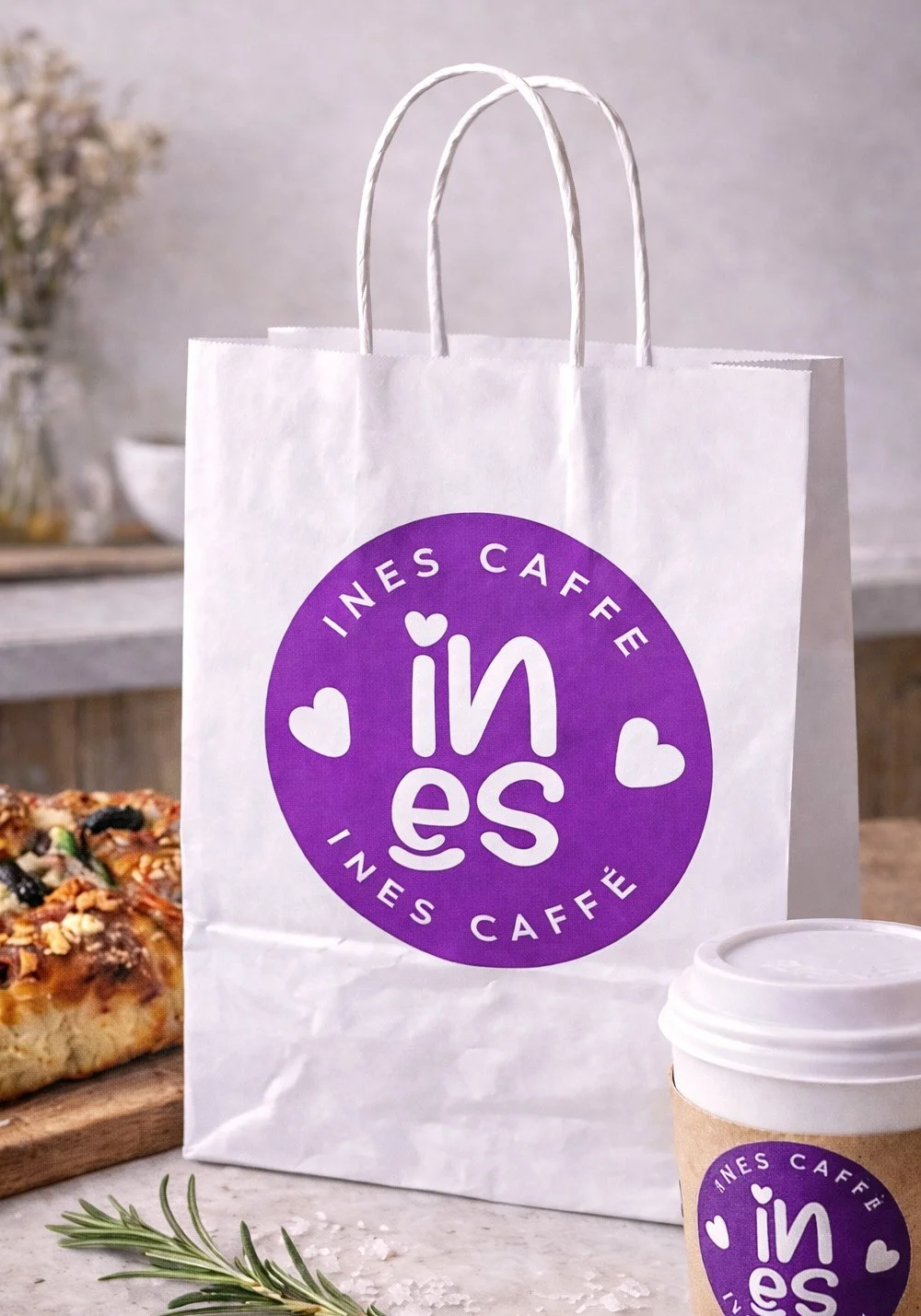 Ines Caffe takeaway bag mockup featuring a white branded paper bag with purple café logo, coffee cup and focaccia in a café setting.