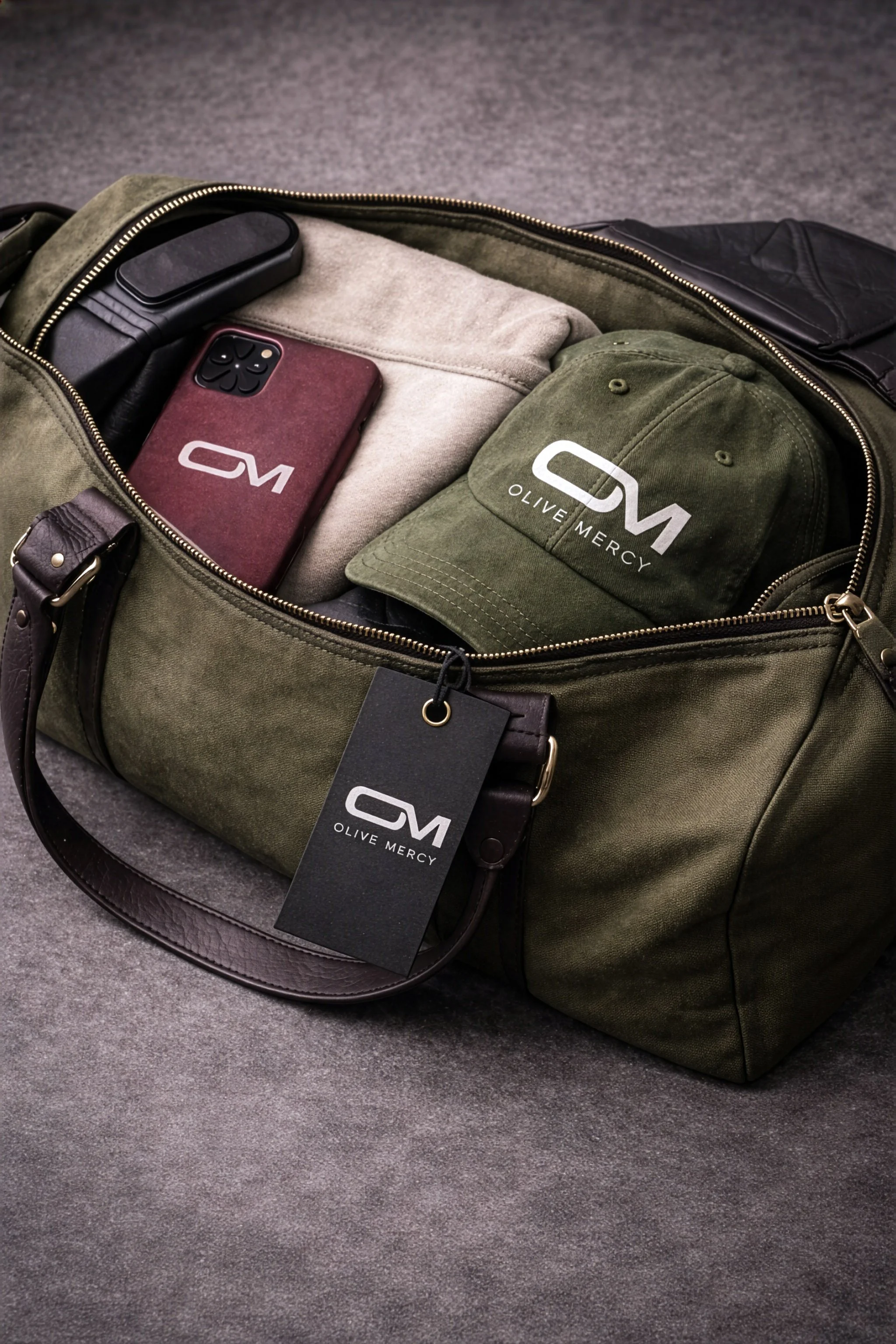 Olive Mercy branded lifestyle mockup featuring an olive bag, cap, swing tag and phone case with the OM monogram logo.