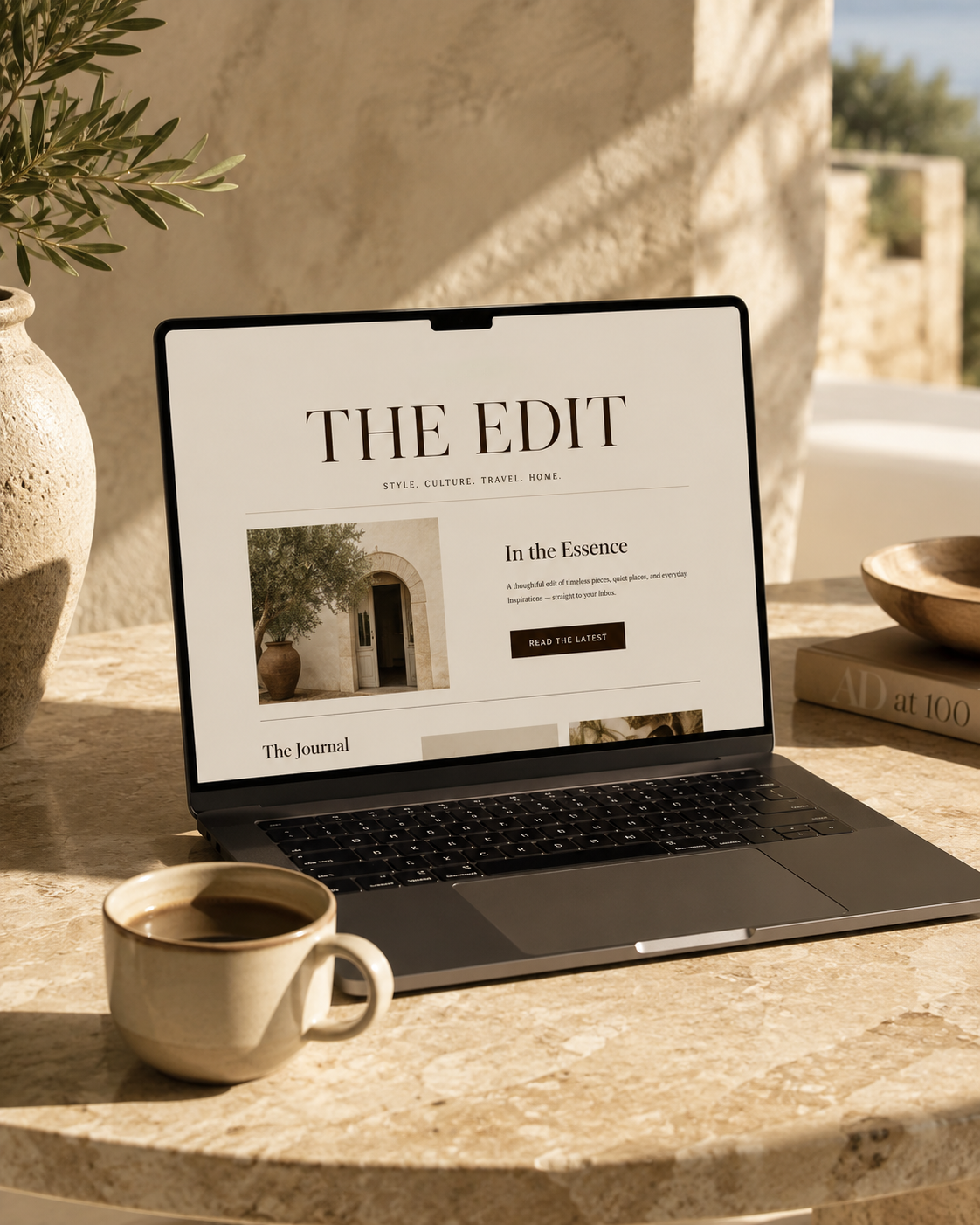 Laptop on a stone table displaying a website titled 'The Edit' with a photograph of an arched doorway and a potted plant inside, with a cup of coffee and a book nearby, in a sunlit outdoor setting.