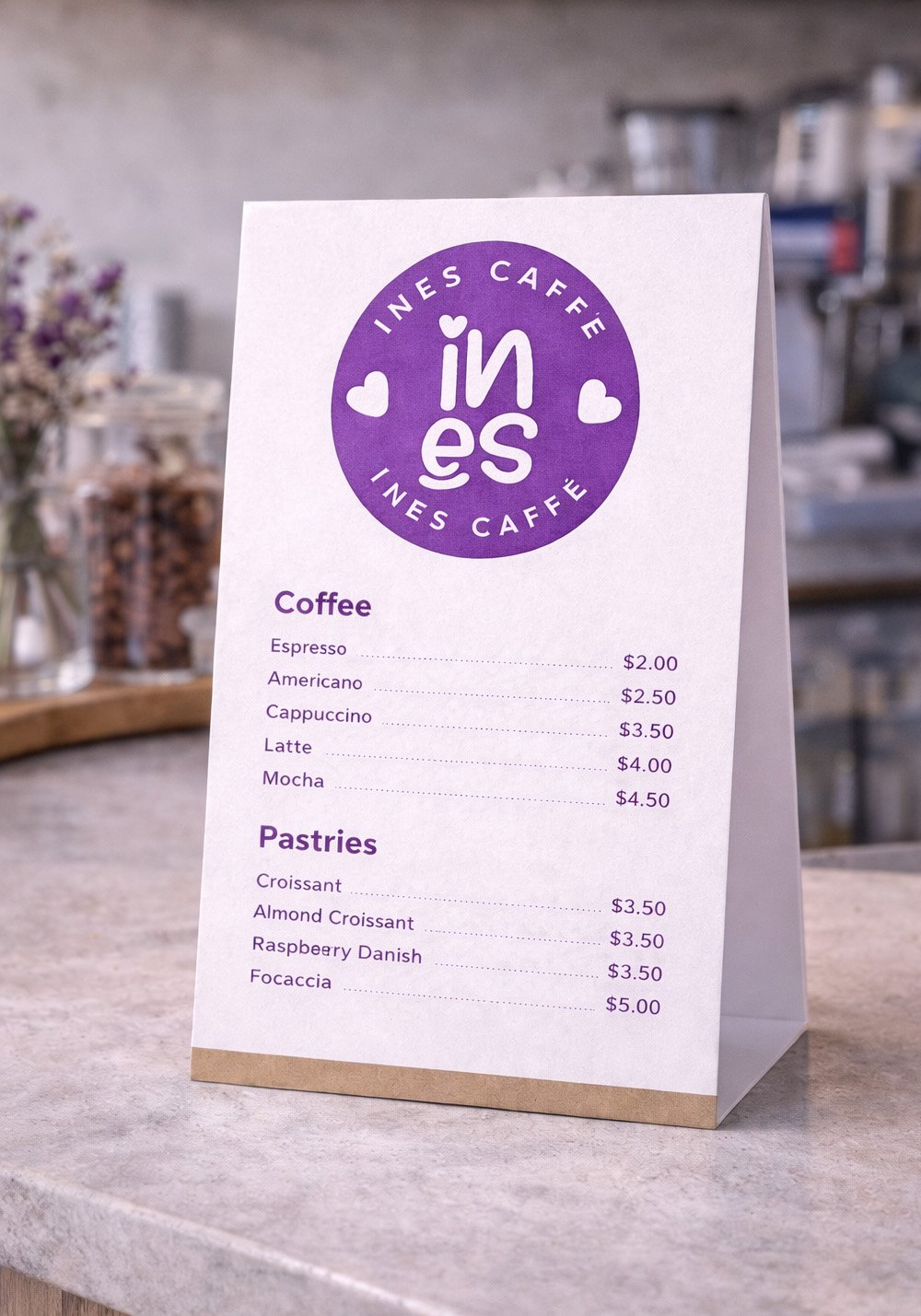 Ines Caffe counter menu mockup featuring a purple café logo, coffee and pastry menu items on a folded tabletop display in a café setting.