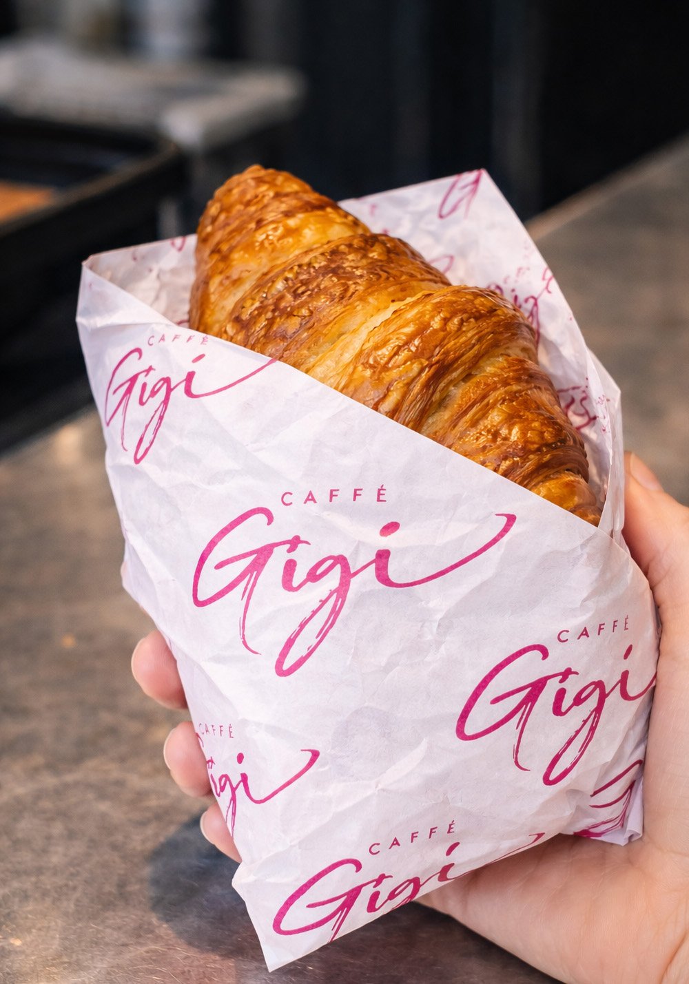 Caffè Gigi croissant wrapping paper mockup featuring a fresh croissant wrapped in branded white tissue paper with pink logo design.