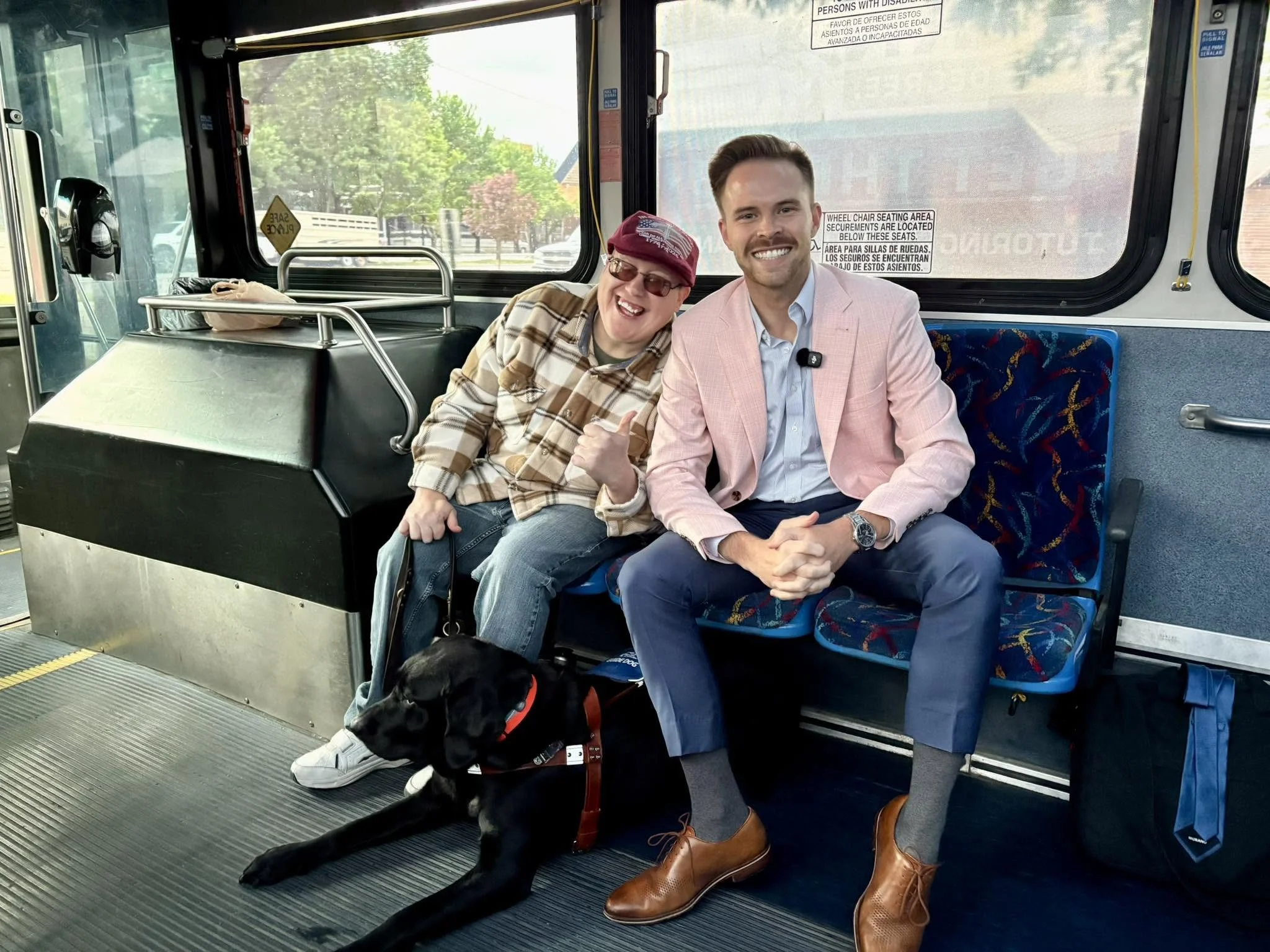 Two men sitting on a bus, smiling at the camera. The man on the left wears a red cap and glasses, with a plaid shirt. The man on the right wears a pink blazer, blue shirt, and brown shoes. A black dog lies at their feet, and the bus interior has larg