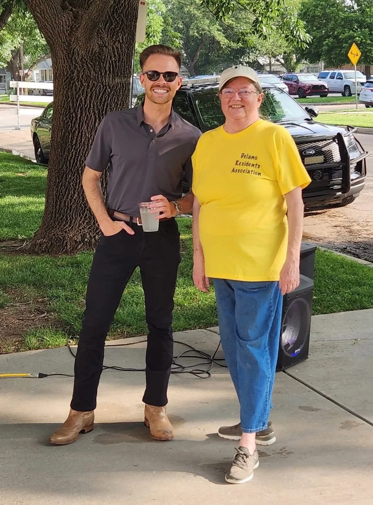 Two men standing outdoors near a large tree. One man is wearing a gray shirt, black pants, tan boots, and sunglasses, holding a clear cup. The other man is wearing a yellow T-shirt with black text, blue jeans, gray sneakers, and a beige cap. There is