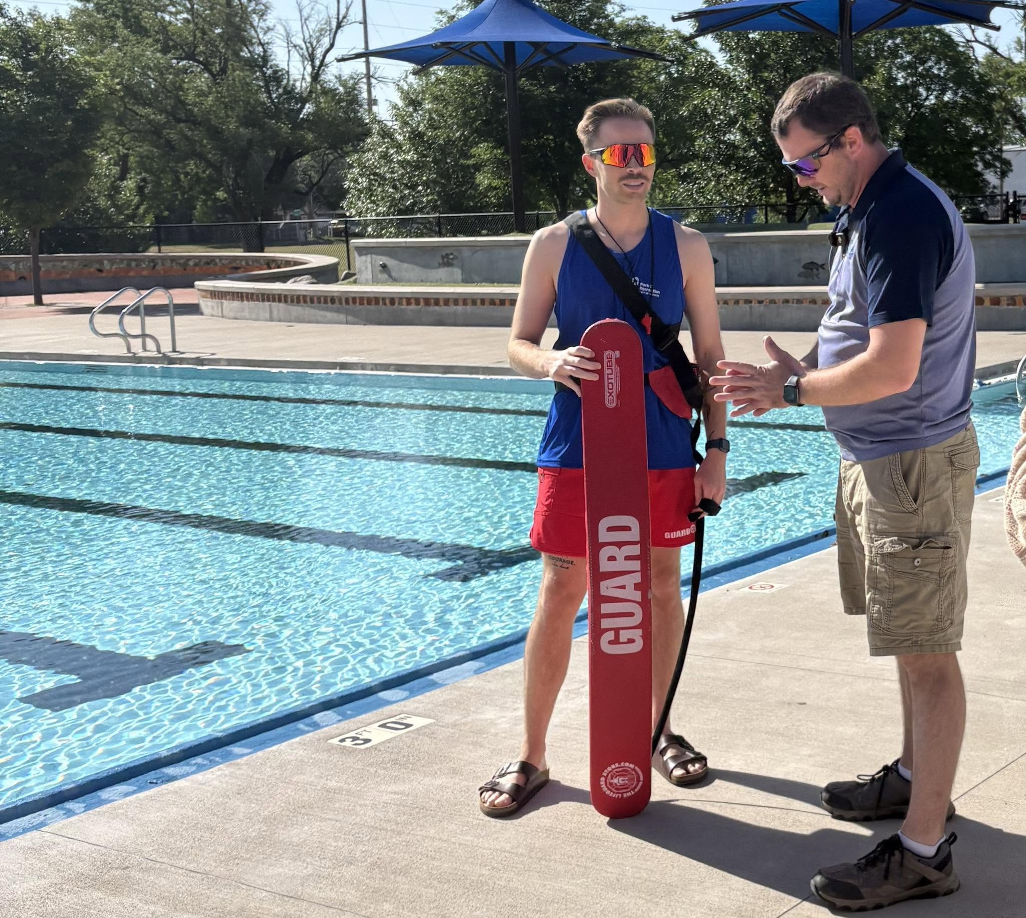 A man in sunglasses and athletic clothing standing by a swimming pool, holding a red rescue float, while another person talks to him. There are blue umbrellas and trees in the background.