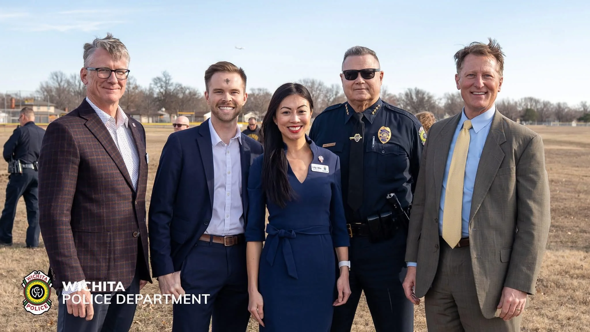 Group of five people standing outdoors at a park, with some police officers and a dog in the background, during daytime, smiling for the camera. The group includes men and women dressed in formal and police uniforms.