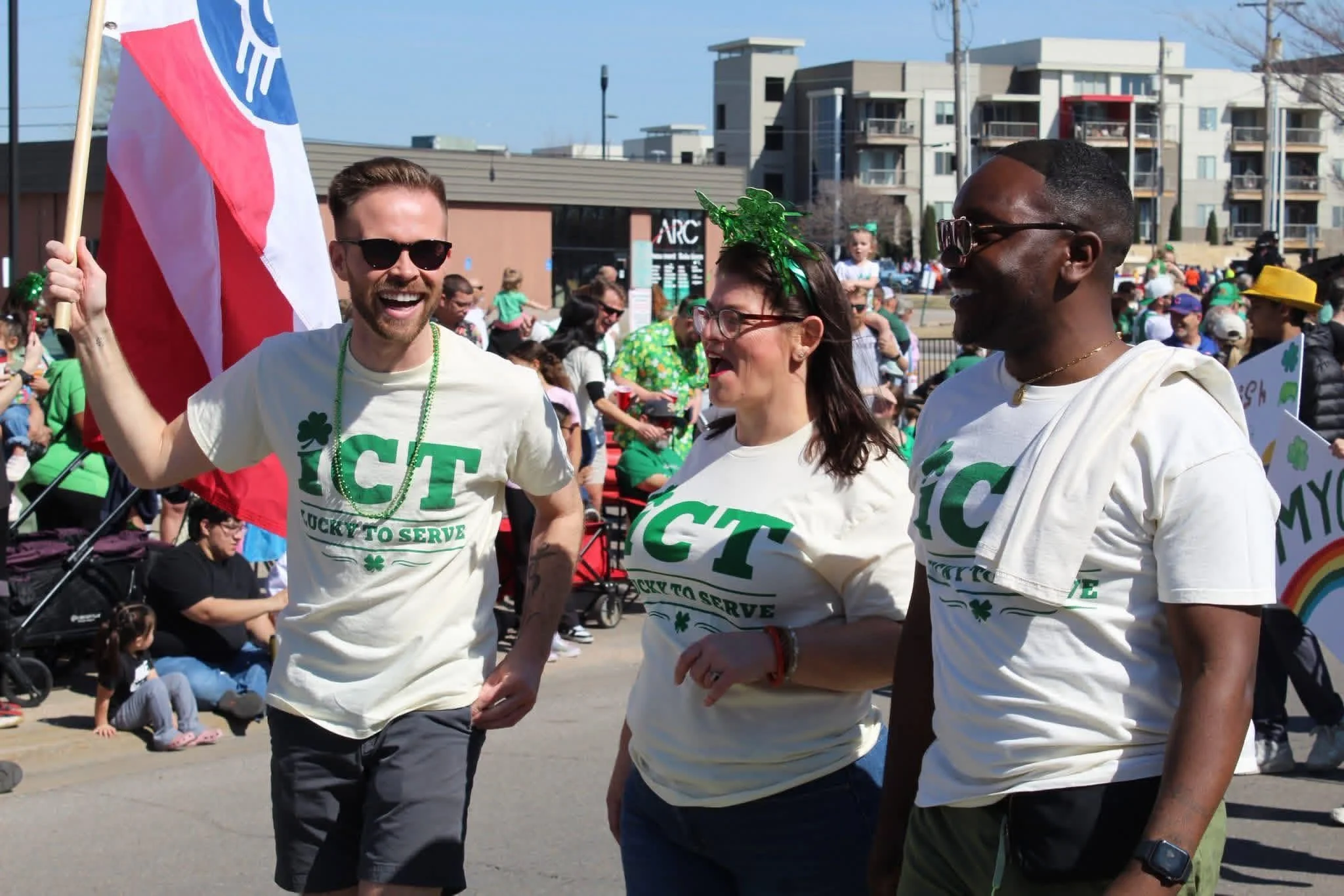 Three people dressed in T-shirts that say "I ♥ CT" and "Lucky to Serve," walking and holding hands, celebrating during a parade or event with many people in the background and some wearing green accessories.